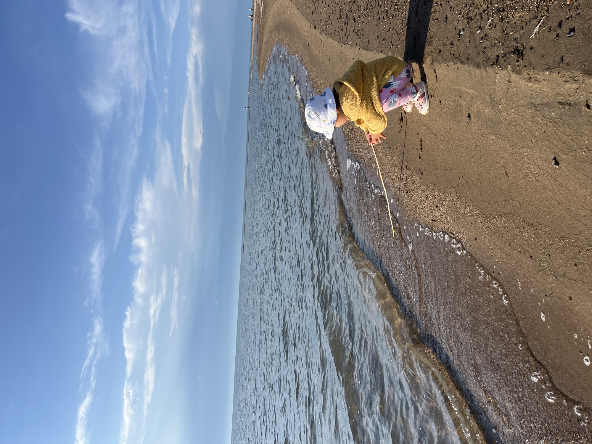 The media file shows a young child playing on a sandy beach next to a body of water under a bright blue sky with wispy white clouds. The child is wearing a yellow sweater, patterned leggings, and a white sun hat, and is crouching down, reaching towards the water with a stick. The scene appears to be set during the daytime, with the sun casting shadows on the sand. The overall mood seems peaceful and contemplative, with the child engrossed in exploring the shoreline. The location is identified as Ak Dobo, Kyrgyzstan, suggesting a natural landscape with a beach area. There is no visible text in the media file.