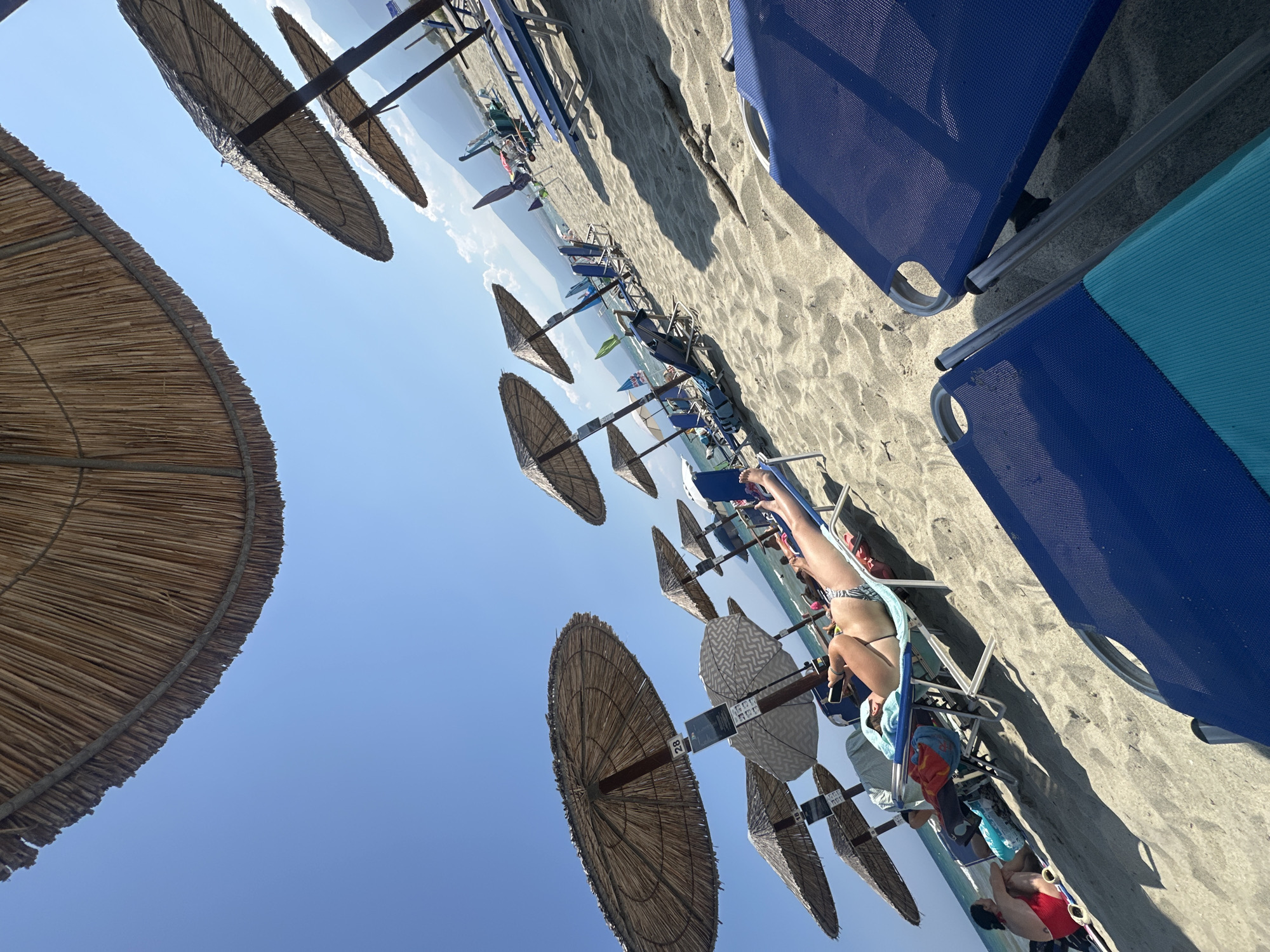 The image depicts a sunny day at a sandy beach in Greece. Several straw umbrellas are erected over empty and occupied lounge chairs, providing shade to beachgoers. In the foreground, two blue lounge chairs are prominently featured, with one partially obscured. A woman is seen relaxing on a lounge chair under an umbrella, holding a smartphone, while another person in red swimwear sits on the sand in the middle ground. In the background, the sea is visible with some beach structures and mountains in the distance under a clear blue sky. The overall scene is one of leisure and relaxation on a summer day.