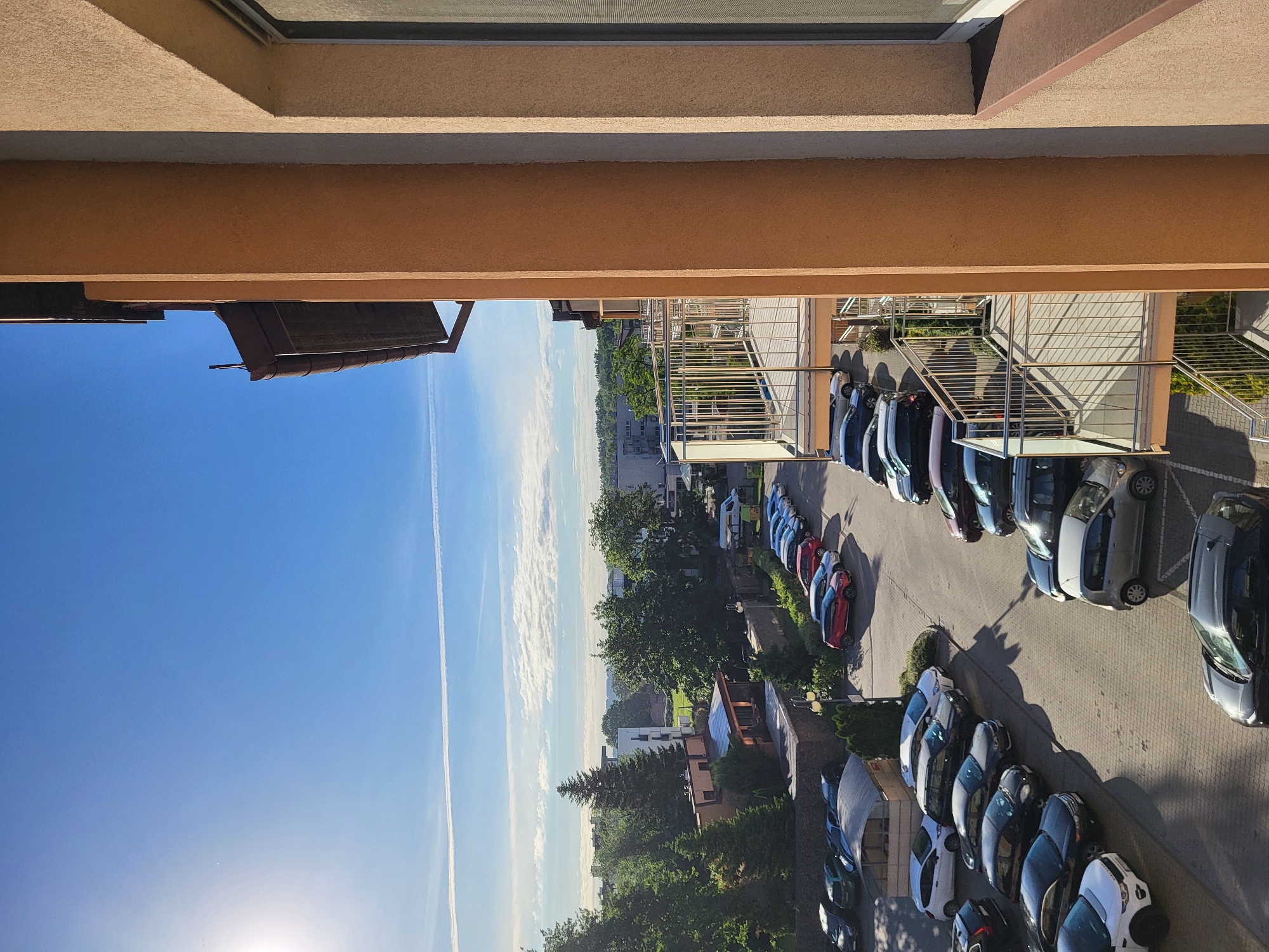 The image captures a high-angle view of a parking area filled with cars of various colors and models. The scene is partially framed by elements of a building, with a light brown architectural structure visible at the top. In the background, residential buildings and green spaces are visible under a blue sky with contrails.