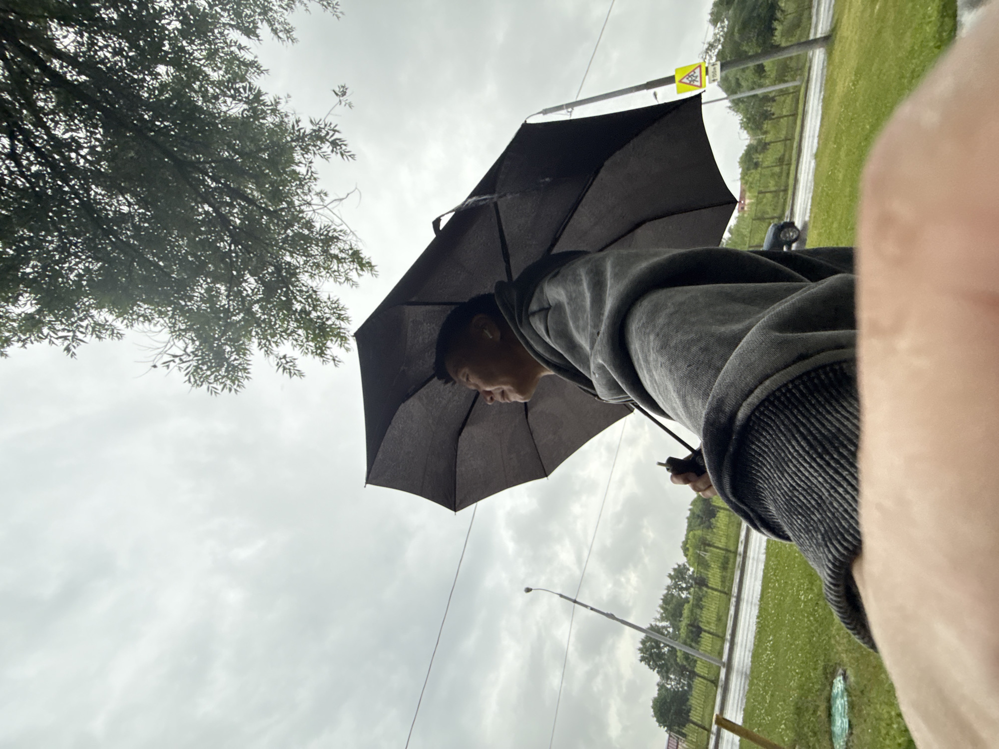 An eye-level, low-angle, outdoor shot captures a person holding a black umbrella on a cloudy day. The person, appearing to be of Asian descent, is wearing a grey hooded sweatshirt. The umbrella is open, shielding the individual from the elements. In the background, a street with a wet road and green grass verge suggests recent rain. Trees and buildings are visible in the distance, hinting at an urban or suburban setting. A street light and a yellow traffic sign with a children crossing symbol are also present. The foreground is dominated by a blurred portion of the person's arm and possibly their leg, suggesting a selfie-style shot. The overall mood is somber due to the overcast sky and wet conditions.