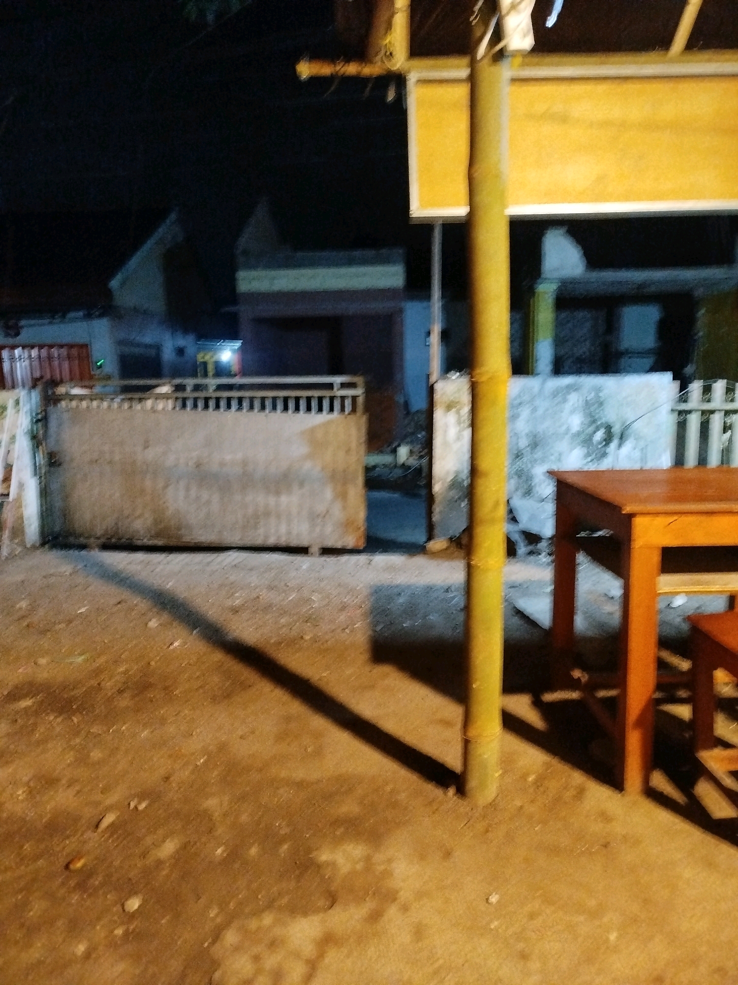 This image depicts an outdoor scene at night, likely in Mojopanggung, Indonesia. In the foreground, a portion of an orange wooden table and a stool are visible on a concrete ground. A thick, yellow bamboo pole stands vertically in the middle ground, casting a shadow. Behind the bamboo pole, a large, weathered metal gate with vertical slats occupies the left side of the frame. Beyond the gate, a dimly lit building with a red-tiled roof and a brown facade is partially visible. The background consists of dark structures, suggesting a residential or commercial area at night. The lighting is artificial and casts long shadows. There are no people present in the image.