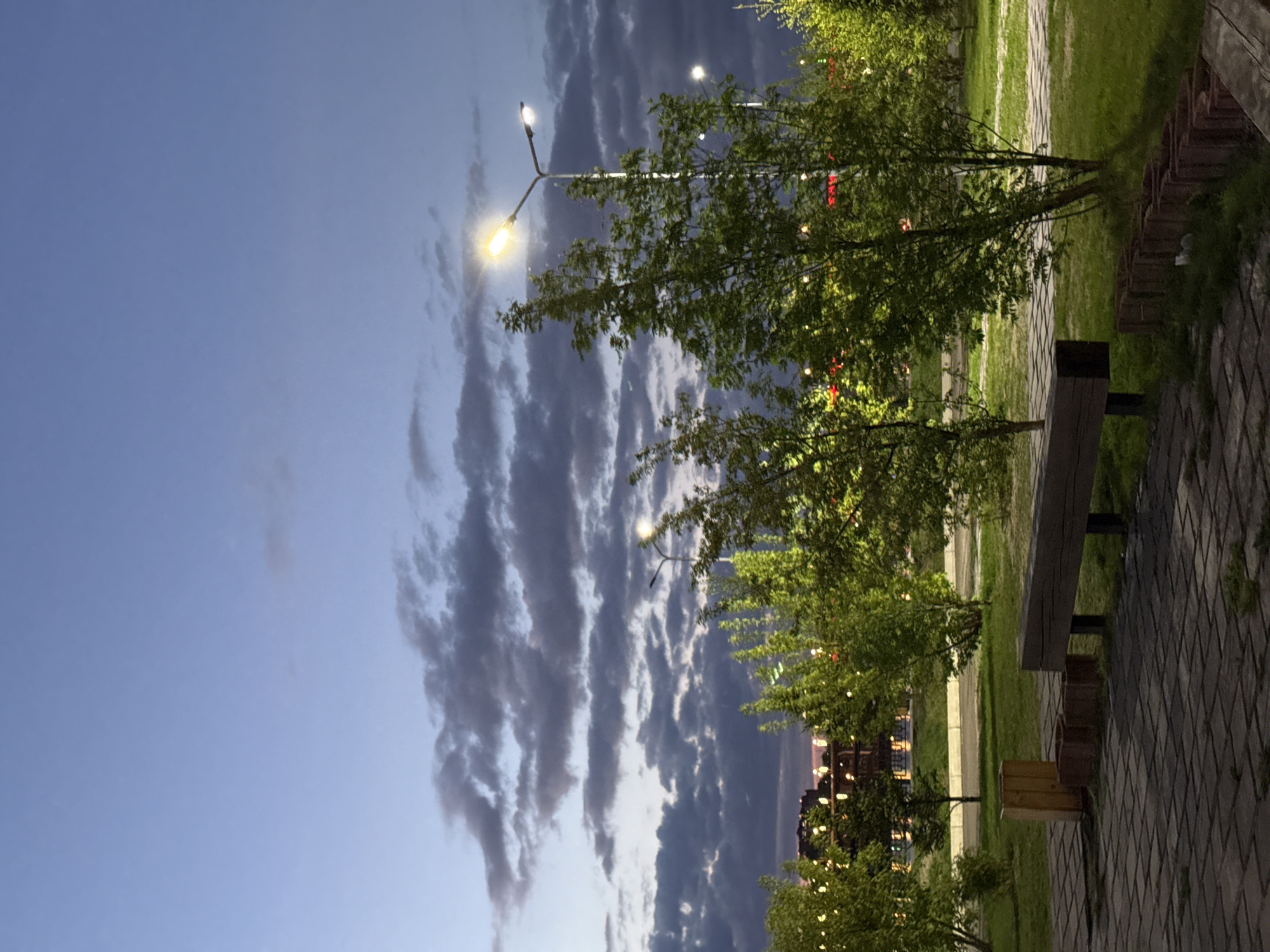 The photograph captures an evening scene in Krasnoyarsk, Russia. A park setting is evident, featuring green grass, trees, and a brick walkway with a bench. The sky is a gradient of blue, with elongated, dark clouds. Streetlights illuminate the area, and building lights are visible in the distance. The image orientation is rotated 90 degrees clockwise.