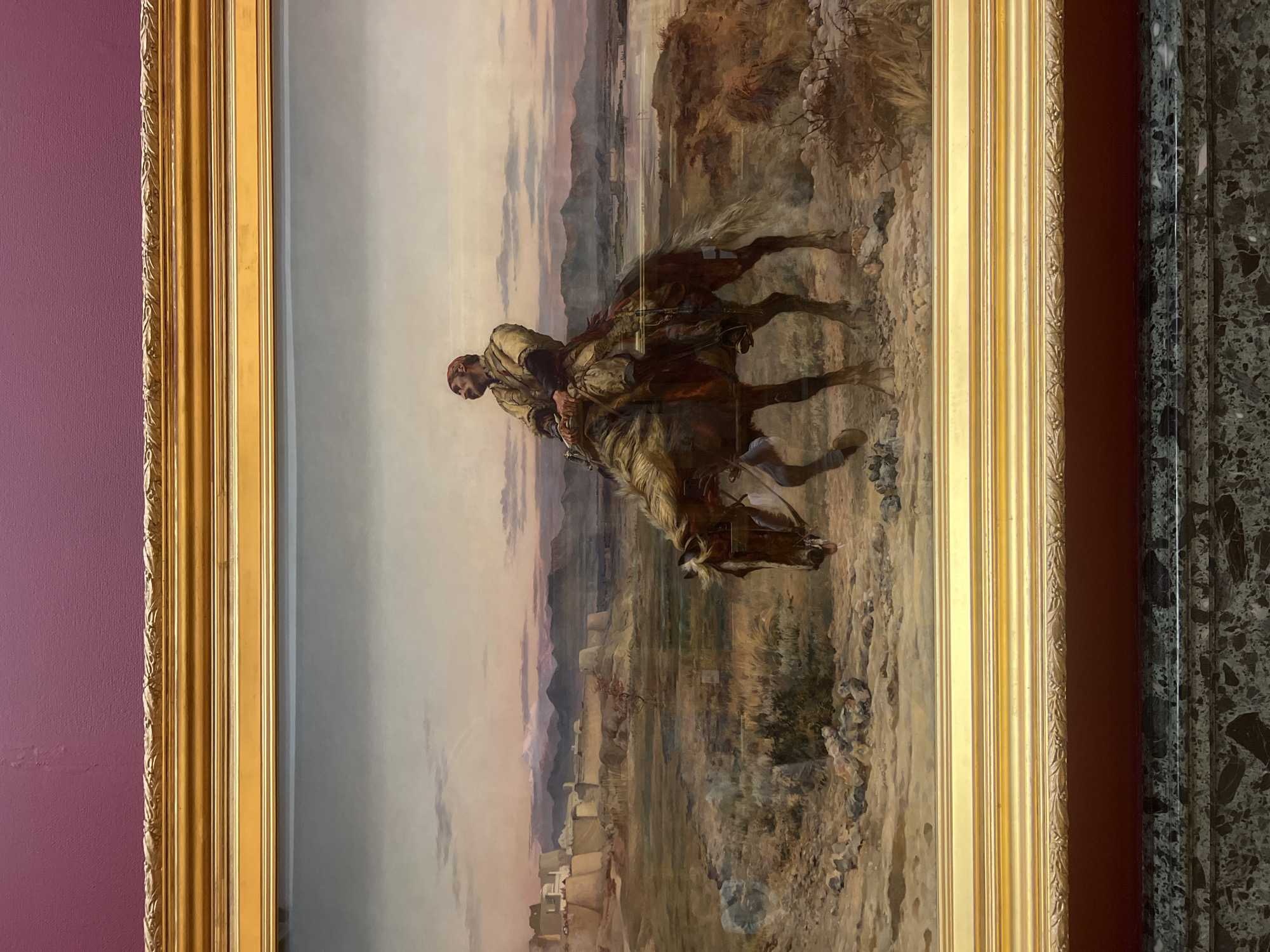 The image displays a vertical painting depicting a solitary man riding a horse through an expansive, arid landscape. The man, positioned in the center-left, is seen in profile, facing right. He wears traditional attire, including a reddish head covering, a light-colored tunic, and layered garments, and has a prominent beard. His light brown and white horse is captured in mid-stride, with a front leg raised, suggesting movement.
The scene is set under a soft, diffused sky with hues of pale blue, pink, and purple, indicating late afternoon or early evening light. In the background, distant mountains rise above what appears to be a scattered settlement of tents or simple structures, suggesting a Middle Eastern or North African setting. The foreground and mid-ground are characterized by dry, rocky terrain with sparse vegetation.
The painting is enclosed within a prominent, ornate golden frame featuring intricate carvings. To the left of the frame, the wall is a textured magenta or deep purple. To the right, a narrow strip of reddish-brown separates the frame from a dark, veined marble panel. No discernible text is visible in the image.