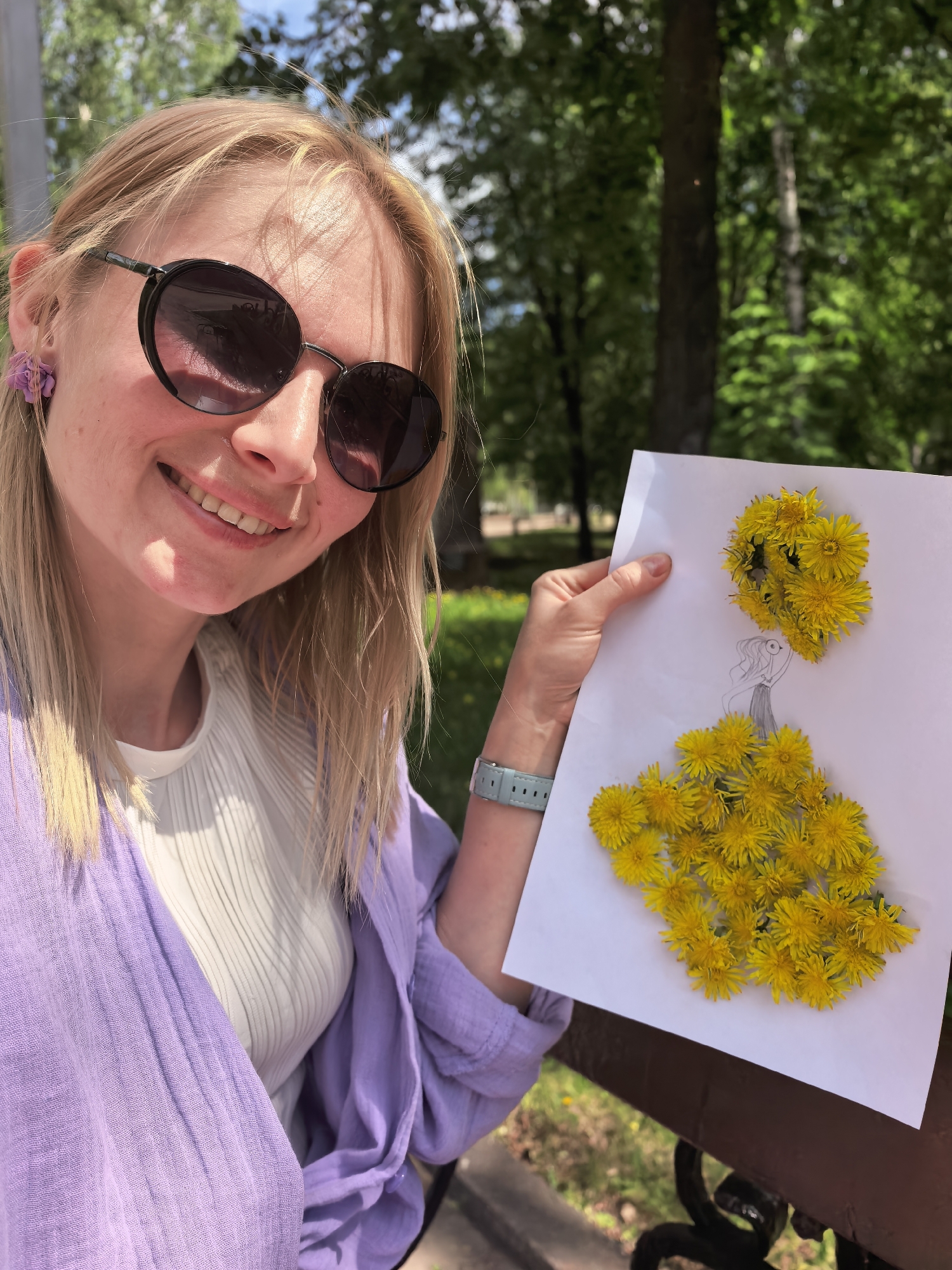 A young, fair-skinned woman with blonde hair smiles directly at the camera while holding up a piece of paper with a drawing of a woman in a dress made of yellow dandelion flowers. She is wearing dark sunglasses, a white shirt, and a light purple, buttoned long-sleeved shirt. In her left ear is a purple flower earring. She has a light blue smartwatch on her left wrist. The paper she holds is on a wooden surface, possibly a park bench. The background shows a green, sunny park with trees. Based on the information available, this photo was taken in Sterlitamak, Russia.