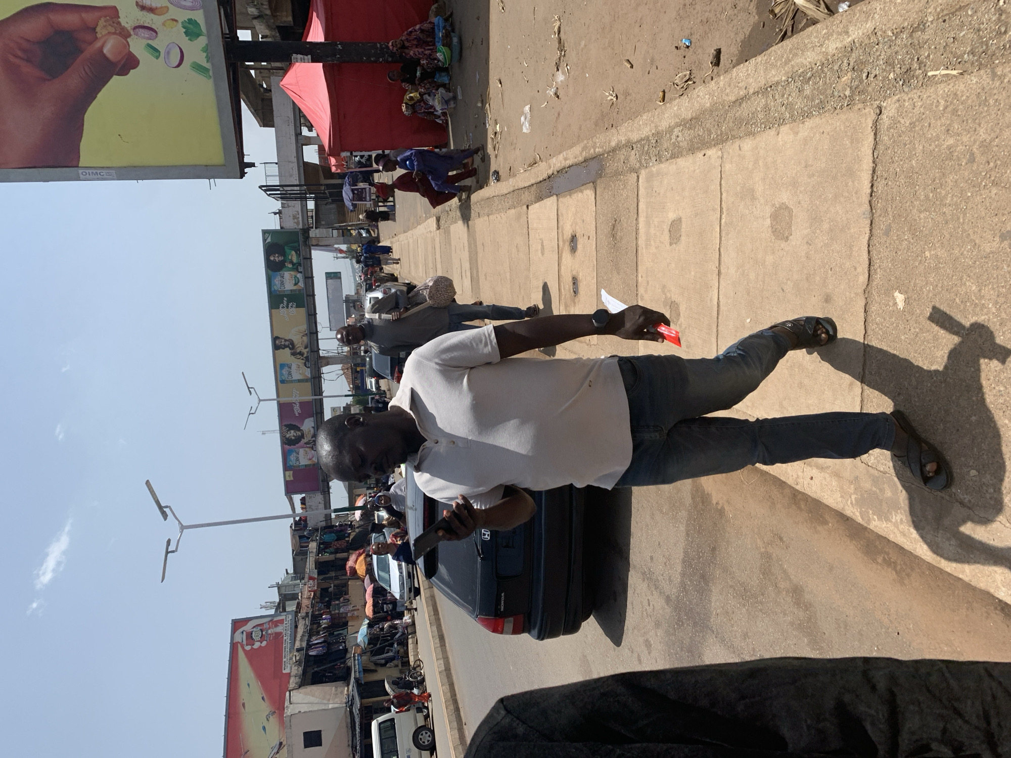 The image depicts a bustling street scene in Jos, Nigeria. The foreground is dominated by a man wearing a white polo shirt and blue jeans, holding a red object and a phone. He appears to be walking on a concrete sidewalk, casting a long shadow due to the bright sunlight. Behind him, a dark-colored Honda car is parked.
The background reveals a busy market environment. Stalls are set up along the street, with various goods displayed. Several people are seen walking, shopping, and interacting within the market. Red tents are visible, suggesting temporary structures for vendors. Further in the background, buildings and billboards are present, including one with a yellow advertisement featuring a hand holding food items. Another billboard shows images of people and text that is partially obscured. Streetlights are also visible. The scene suggests a lively daytime atmosphere, likely mid-morning or afternoon, given the strong shadows and clear sky. The overall impression is one of vibrant daily commerce and activity.