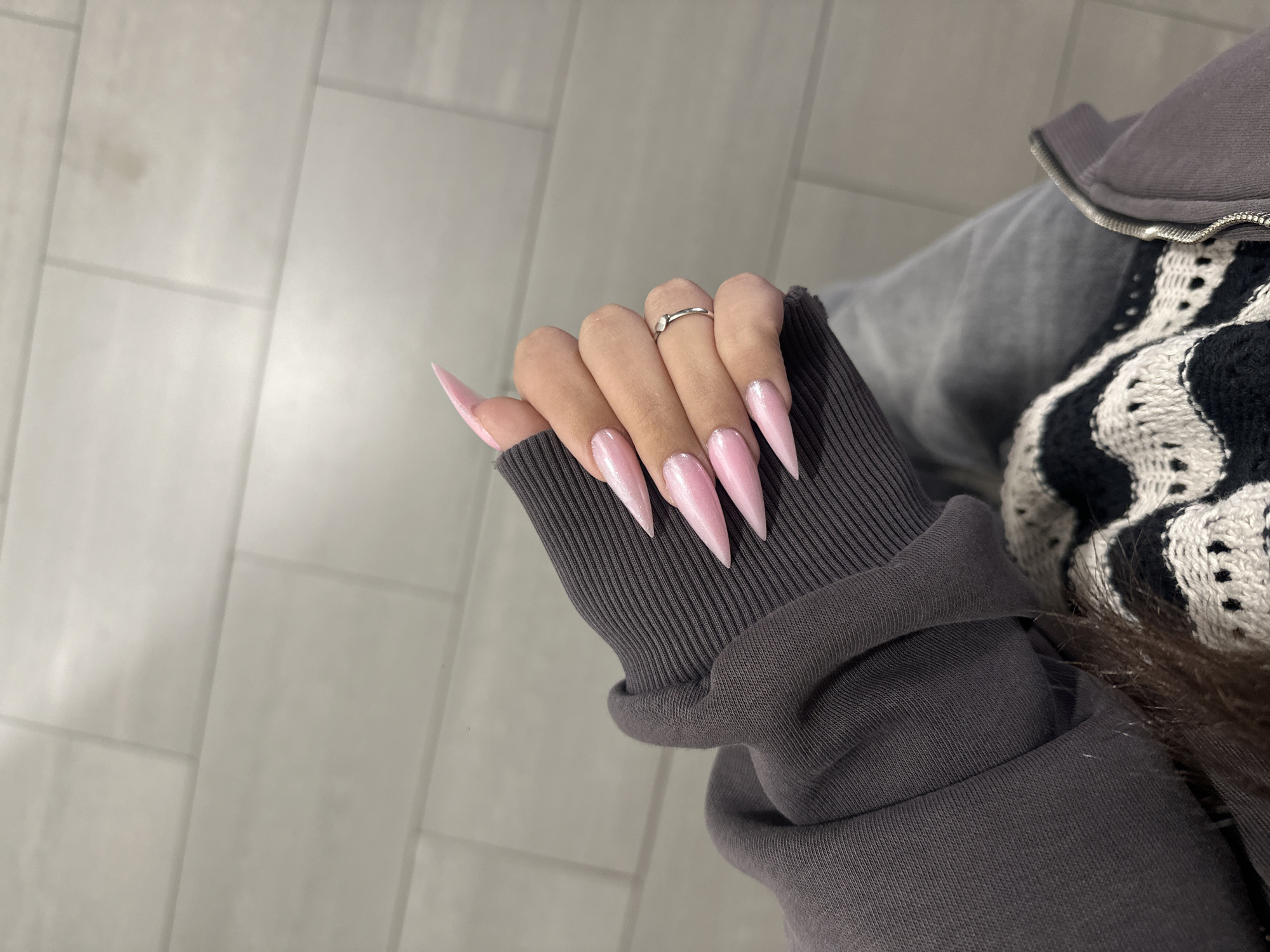A person's hand is displayed, adorned with long, pointed pink nails that catch the light. A simple silver ring is on the middle finger. The hand rests on a gray ribbed sleeve of a sweatshirt or jacket. Adjacent to the sleeve, partially visible, is a black and white patterned sweater. The background is a neutral, light gray tiled floor or wall. The location is unknown and cannot be determined based on the image. Therefore, a specific location within the city of Yerevan, Armenia, cannot be determined.