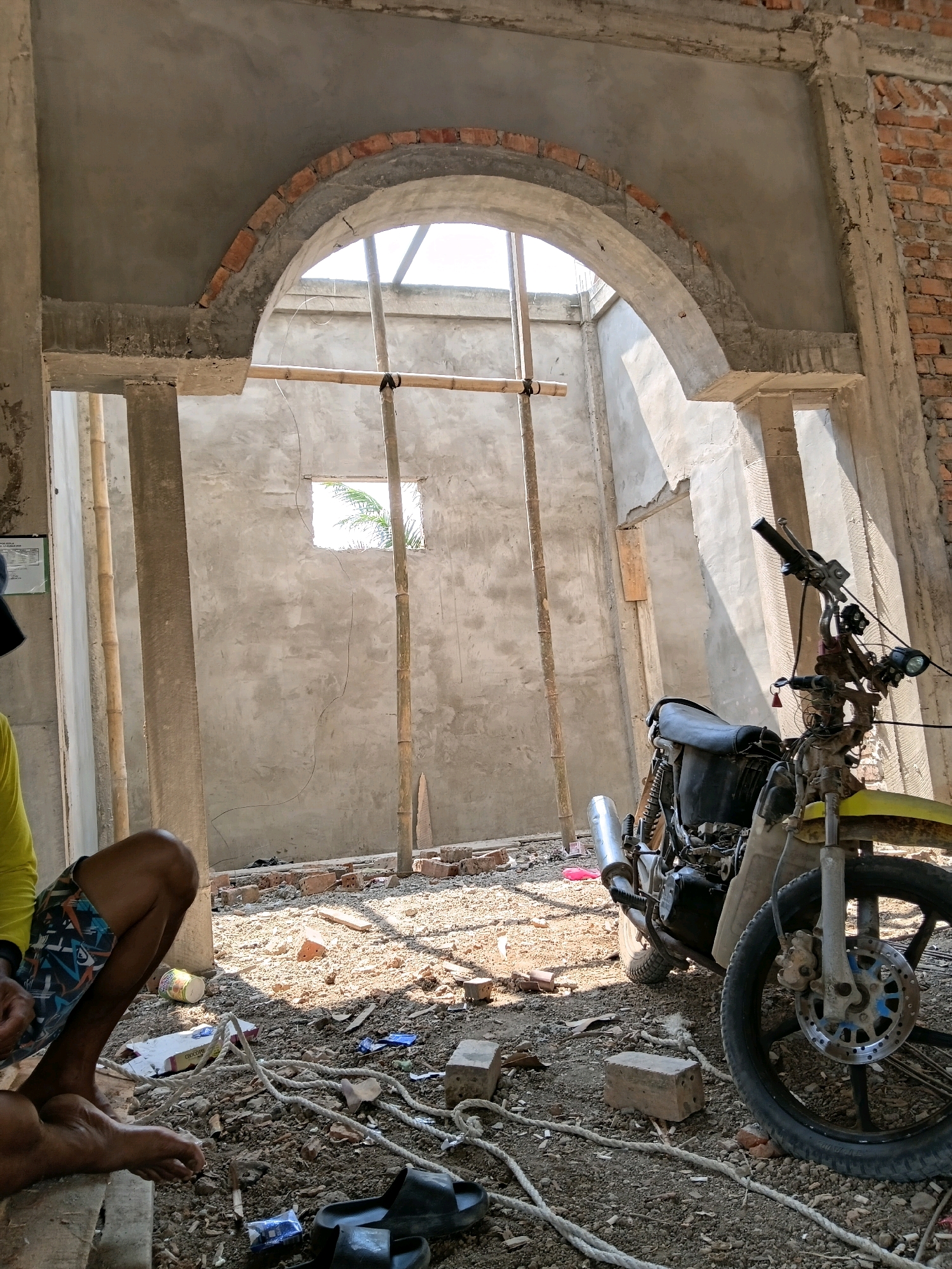 The image depicts an unfinished construction site in Margo Rukun, Indonesia. In the foreground, a person's legs and feet are visible, wearing patterned shorts and sitting on a wooden plank. Next to them, on the dusty ground, are a pair of black sandals, a roll of rope, and some debris, including bricks and discarded packaging.
Dominating the mid-ground is a motorcycle, its front wheel and handlebars prominently featured. The motorcycle appears to be old and somewhat weathered. Behind the motorcycle and the seated individual, the construction site unfolds. An arched opening, partially constructed with brick and concrete, is visible. Bamboo poles are used as temporary supports within the structure.
The walls are made of rough, unfinished concrete, with exposed brickwork visible on the right side. A small, rectangular opening in the back wall allows a glimpse of palm fronds and sunlight. The ground is covered with loose soil, gravel, bricks, and scattered construction materials. Sunlight casts strong shadows across the scene, indicating it is likely daytime. The overall impression is one of ongoing construction and a utilitarian, somewhat unkempt environment.