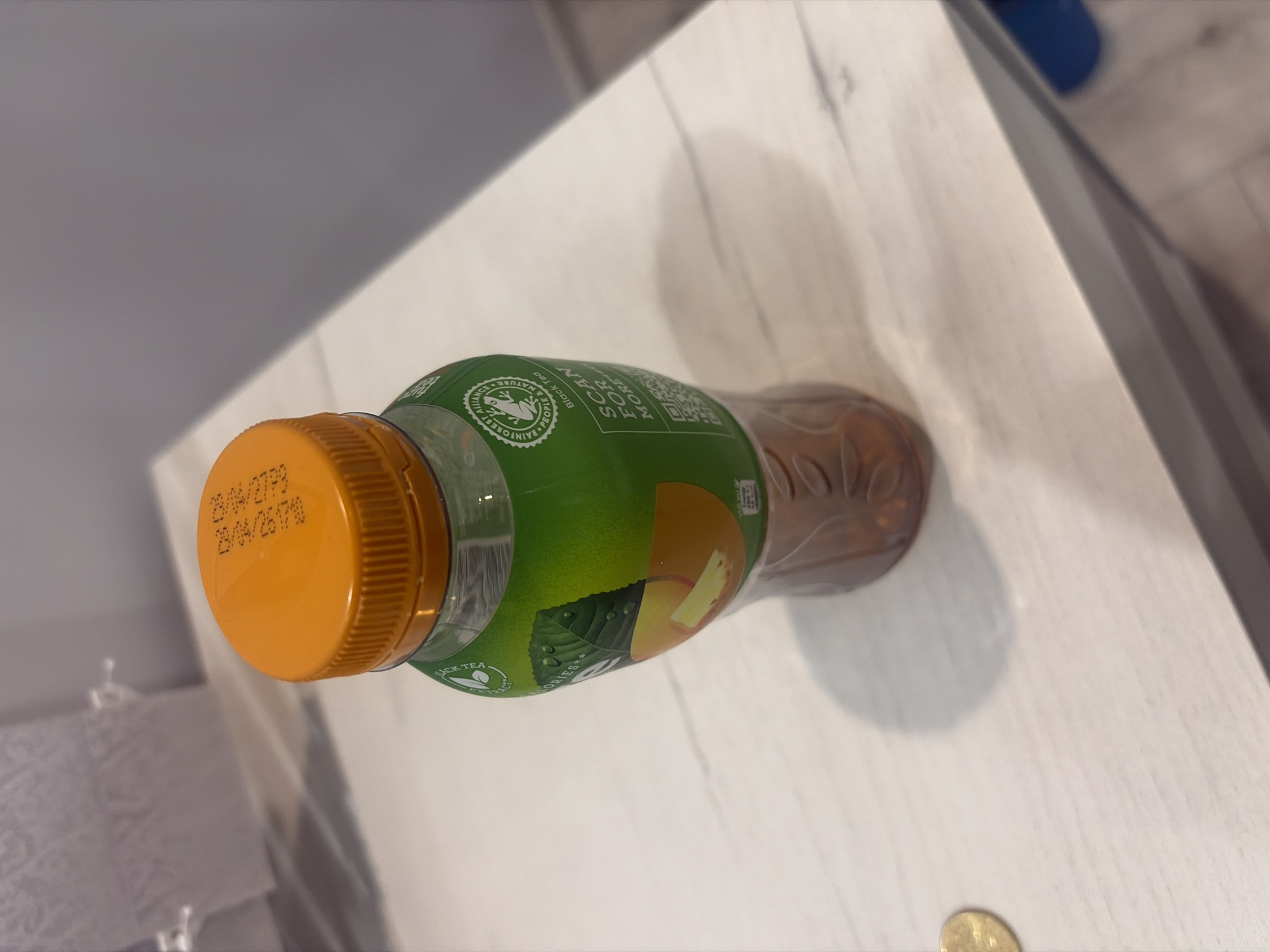 A close-up, slightly high-angle shot shows a bottle of iced tea with a bright orange cap on a light-colored wooden surface. The label is predominantly green with an image of peach slices and leaves. Text on the label includes "SCAN FOR MORE" and a QR code, along with a circular logo indicating "RAINFOREST ALLIANCE CERTIFIED PEOPLE & NATURE BLACK TEA". Embossed text, likely a manufacturing or expiry date, is visible on the orange cap. The bottle contains a brownish liquid, and the texture of the wooden surface, which appears to be a tabletop or shelf, is subtly visible. The background is out of focus, showing what appears to be a roll of toilet paper on the left and a gray wall on the right. A small, golden coin is also present on the surface to the left of the bottle. The scene is static and appears to be indoors, likely in a bathroom or kitchen area, given the presence of the toilet paper. The location of Podgumer, Bulgaria, is not depicted in the image.