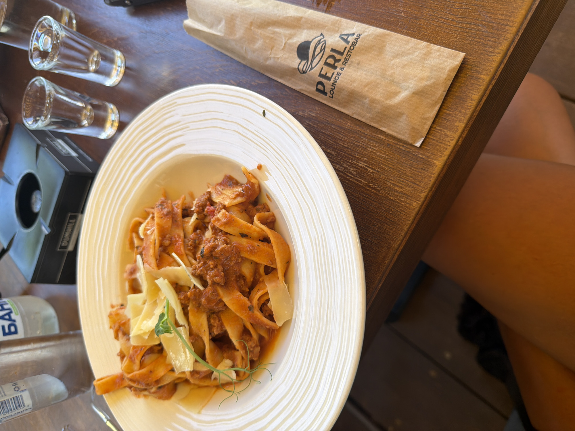 A close-up, overhead shot shows a plate of pasta with a rich red sauce and shaved cheese on a rustic wooden table. To the right of the pasta, a brown paper bag with the logo "PERLA LOUNGE & RESTOBAR" is visible. In the background, out of focus, are two shot glasses and a bottle of water. The setting appears to be a restaurant in Primorsko, Bulgaria, indicated by the restaurant's name. The scene captures a moment of dining, with the focus on the food and the restaurant's branding.