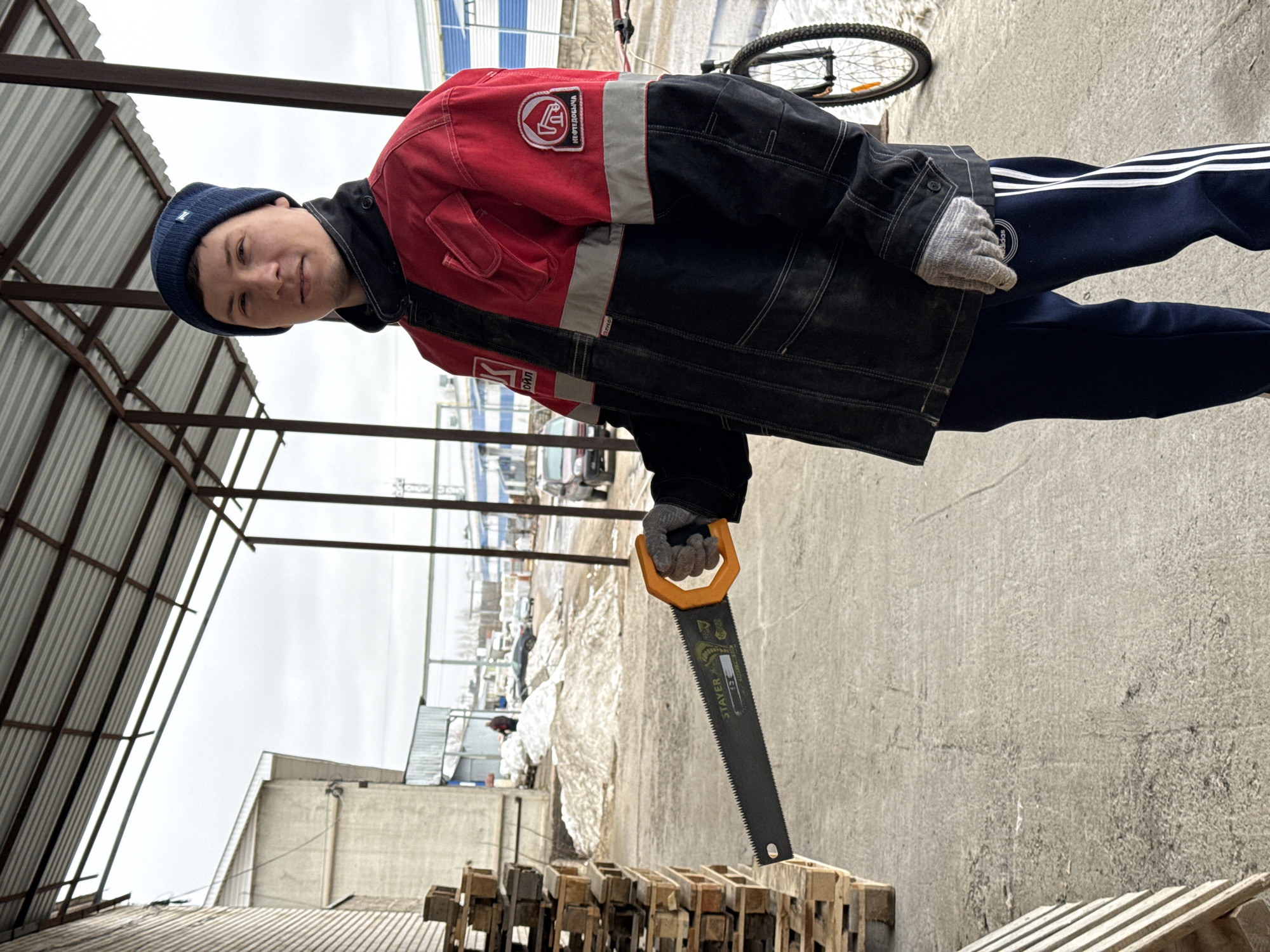 A person wearing a navy blue beanie, a black and red jacket with reflective stripes, and dark blue sweatpants with three white stripes is standing outdoors. They are holding a handsaw with an orange handle over a stack of wooden pallets. The background includes a concrete area, snow-covered ground, a corrugated metal structure, and a bicycle wheel partially visible. The location is Chaikovsky, Russia.