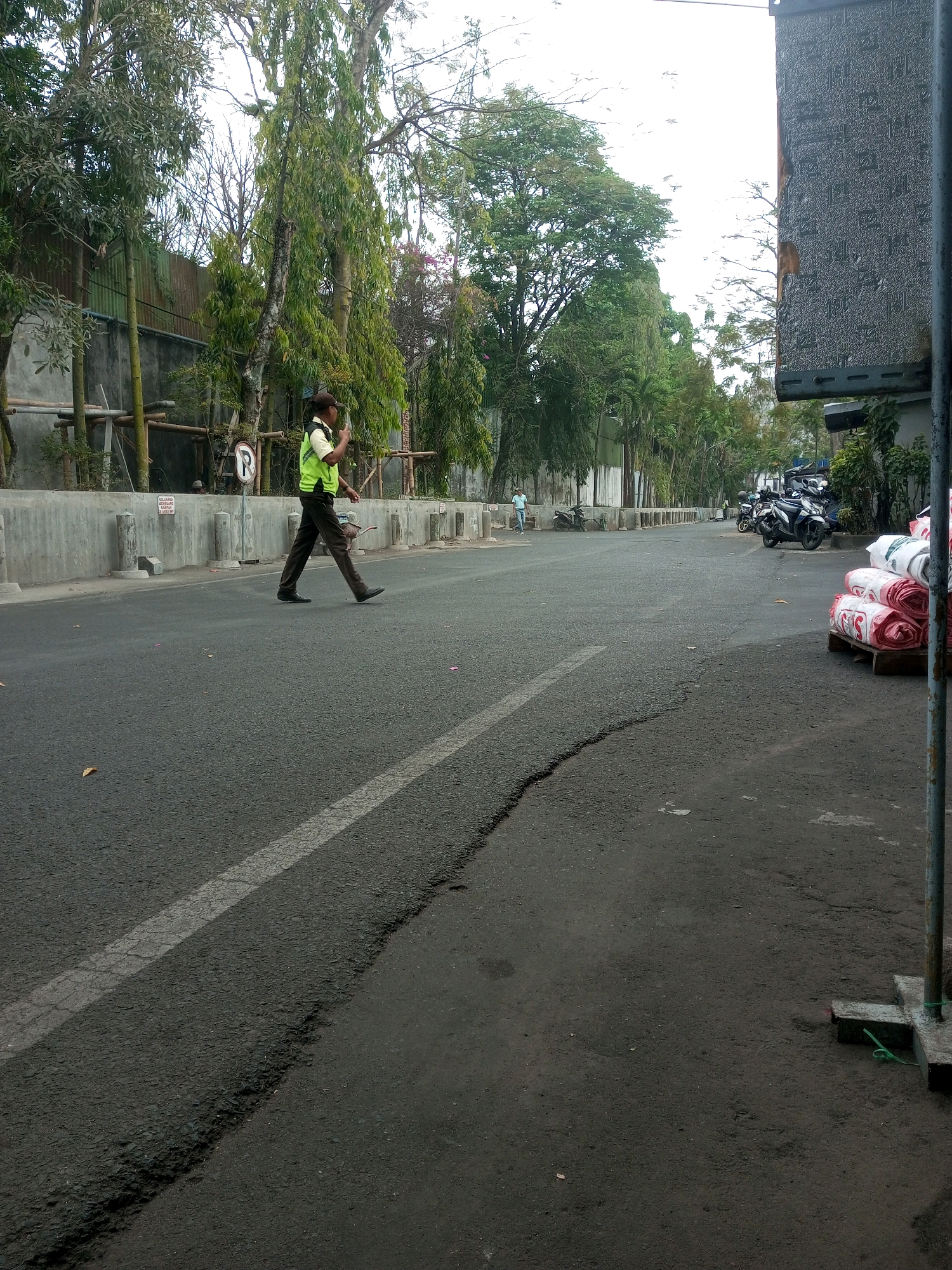 A person wearing a high-visibility vest and dark pants walks across a paved road in Malang, Indonesia. The person is positioned in the left half of the frame, moving from left to right. Behind the person, a concrete barrier runs along the side of the road, with trees and foliage beyond it. A "P" sign is visible on the barrier. In the distance, several motorcycles are parked, and a stack of rolled materials, possibly fabric or carpet, is on a wooden pallet on the right side of the road. The road surface shows some wear and tear, with a prominent crack running diagonally across the foreground. The sky is overcast.