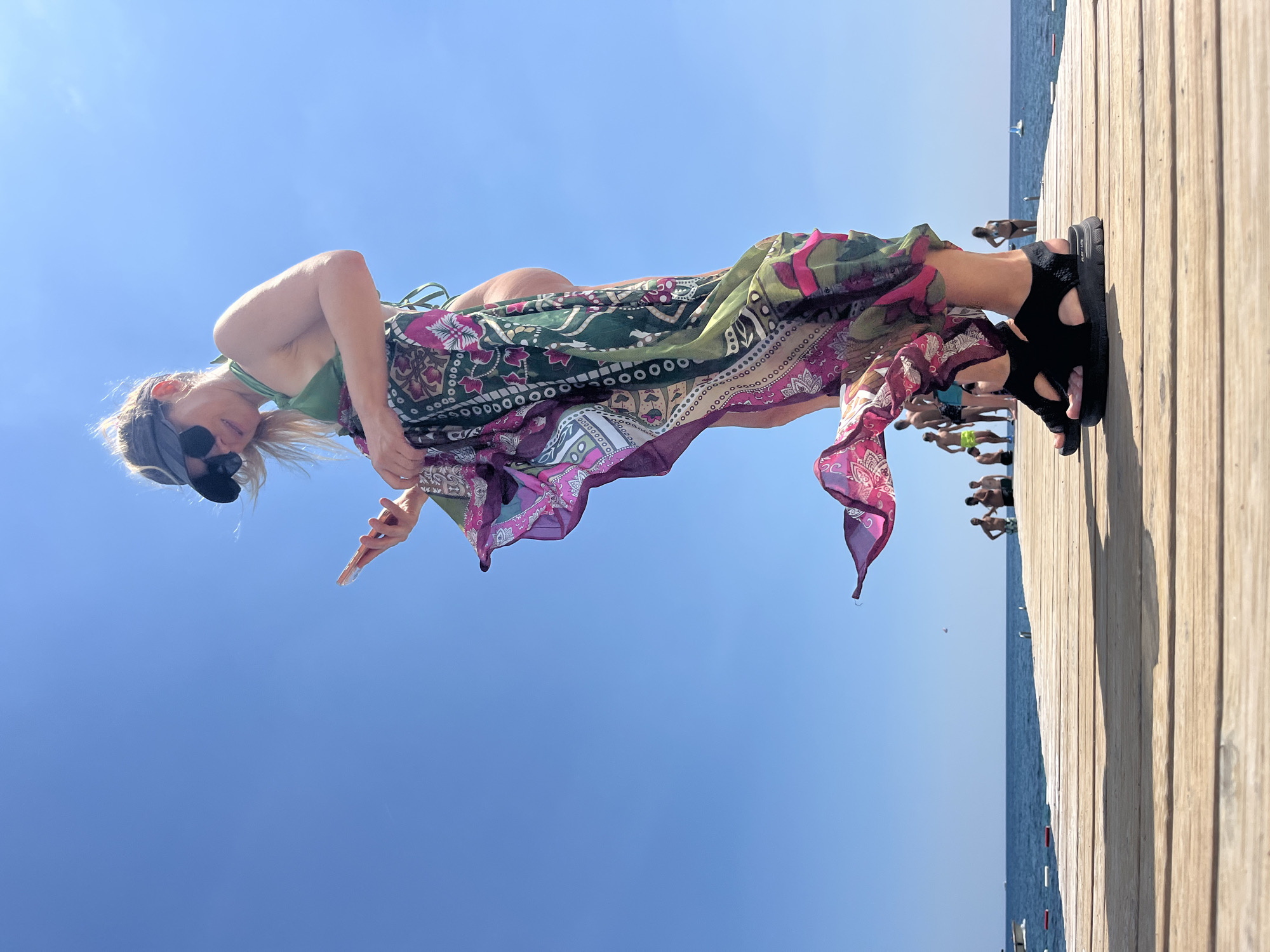 The image features a woman on a wooden pier with a clear blue sky above and the ocean in the background. She is wearing a green bikini top, a colorful patterned sarong, black sunglasses, and black sandals. The sarong is draped over her and flows around her. Her body is positioned in a way that suggests she is looking down and possibly posing.
In the background, other people are visible on the pier and in the water, some swimming or standing. The sea is calm and a light blue. The pier is made of light-colored wooden planks. The sun is bright, indicating it is likely daytime, and the sky is cloudless, suggesting good weather. The overall scene appears to be a beach or resort setting.
The image is oriented vertically, making it appear as if the subjects are on a horizontal plane. The woman's expression is not clearly discernible due to the sunglasses and her pose. The setting evokes a sense of leisure and vacation. There is some text visible on the woman's sandal that reads "FLEX-LITE".
