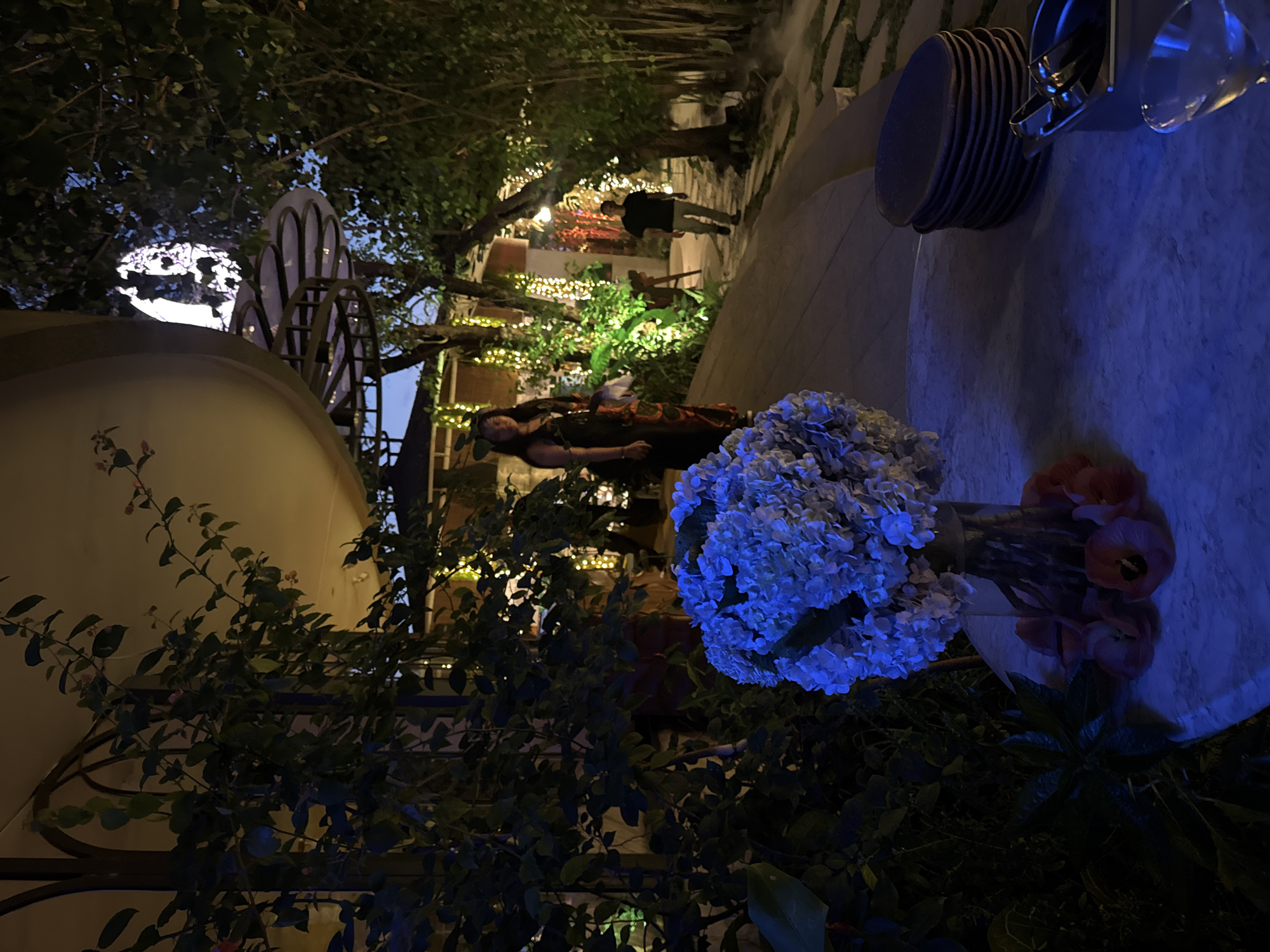 The image depicts an outdoor evening scene at a restaurant in North Kuta, Indonesia. A woman in a patterned dress stands on a pathway, while a man in a black t-shirt walks away from the camera in the background. In the foreground, a table is set with a vase of blue hydrangeas and a stack of dark plates. The restaurant is decorated with numerous fairy lights, illuminating the lush greenery surrounding the area. The overall ambiance suggests a tranquil and inviting atmosphere.