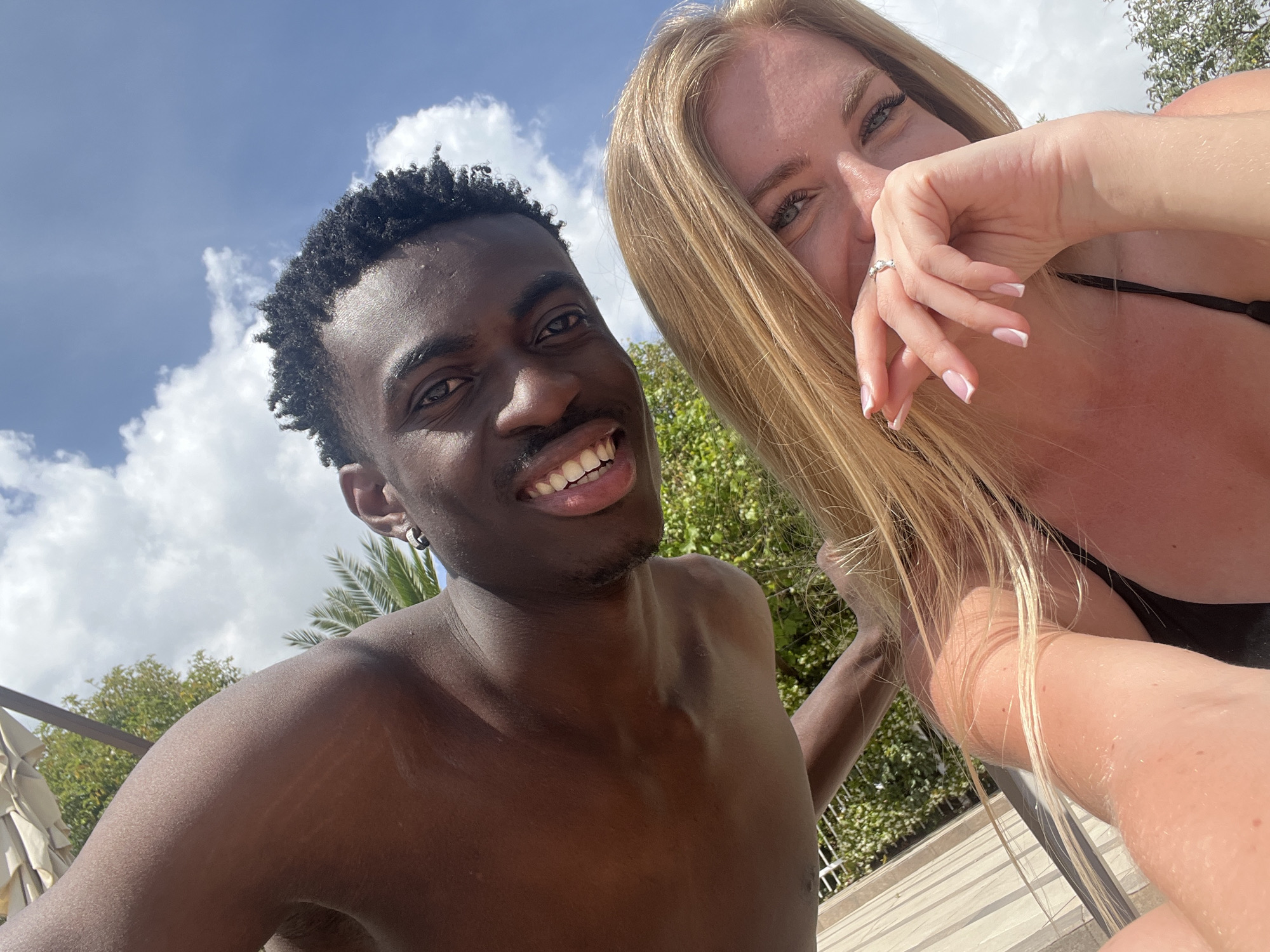 A selfie of a smiling man and a woman with long blonde hair in an outdoor setting. The man is dark-skinned, shirtless, and wearing an earring in his left ear. The woman is fair-skinned, wearing a black top, and partially covering her face with her hand, which has a ring on the ring finger and manicured nails. The background includes trees, a blue sky with clouds, and possibly umbrellas or parasols. It appears to be a sunny day. The exact location in Budva, Montenegro, cannot be determined from the image alone.