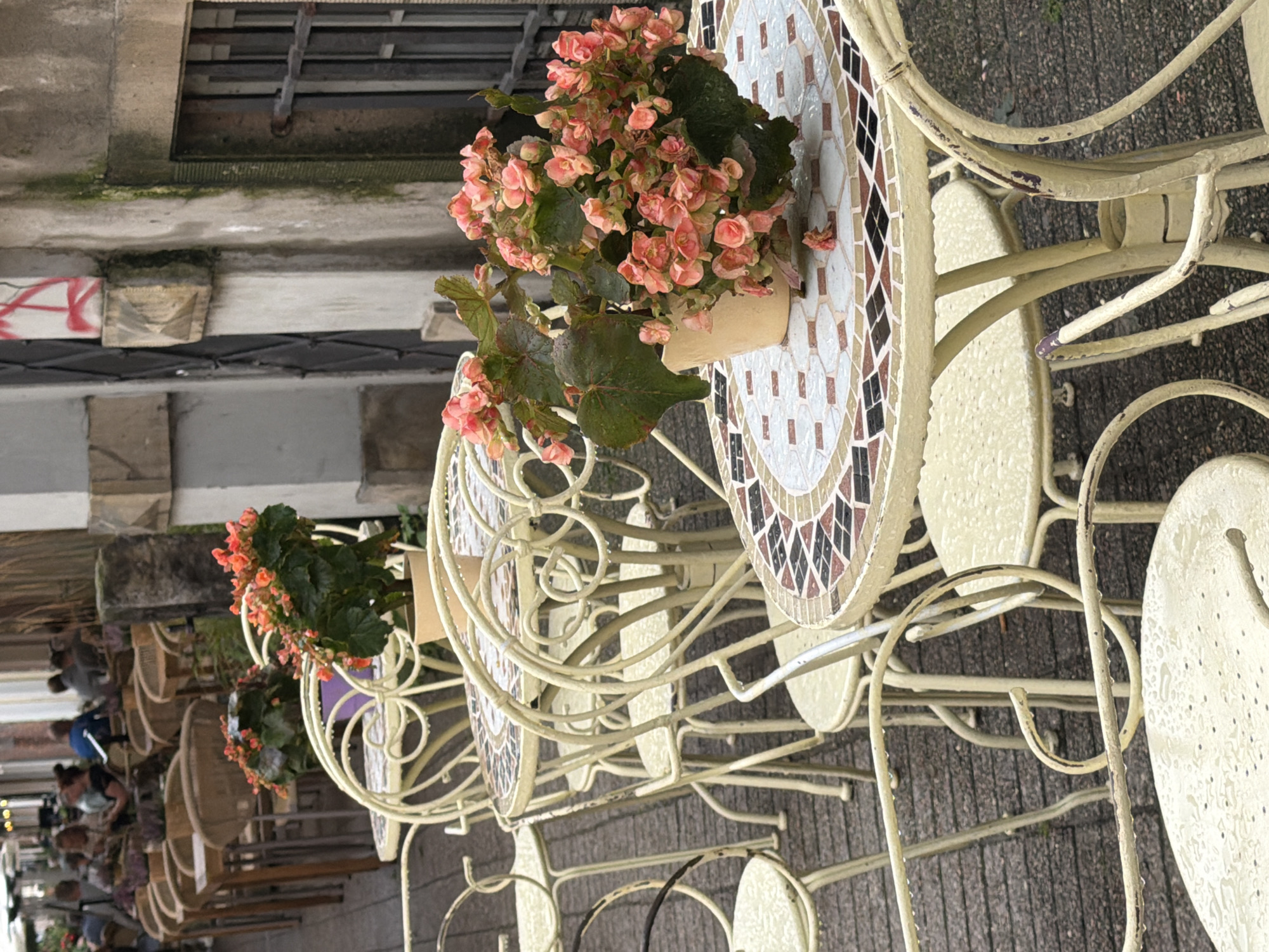 The image depicts an outdoor seating area of a cafe or restaurant. The foreground features several cream-colored metal tables and chairs with intricate scrollwork designs. The tabletops are decorated with mosaic patterns in shades of white, brown, and black. A potted plant with vibrant pink begonia flowers sits on one of the tables. The furniture and pavement are wet, indicating recent rain. In the background, a row of similar tables and chairs are visible, some occupied by patrons. Beyond the seating area, a building with stone and concrete facade is present, featuring a barred window. People are seated at tables, some are conversing, others appear to be looking at their phones or enjoying the ambiance. The scene suggests a European city, potentially Gdańsk, Poland, given the architectural style and the presence of outdoor cafes.