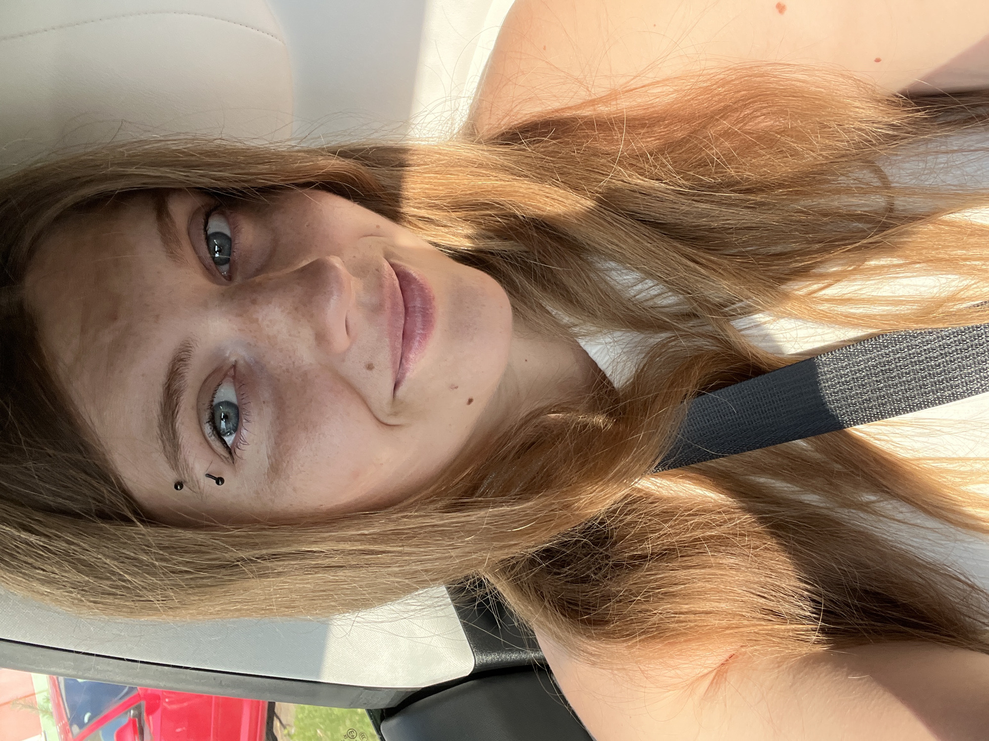 A close-up, front-facing selfie of a young woman with fair skin and light brown hair. She is sitting in the passenger seat of a vehicle, as indicated by the seatbelt across her chest and the car interior visible behind her. A red car is partially visible through the window on the left side of the frame, suggesting an outdoor setting, possibly in a suburban or rural area. The woman has blue eyes, freckles across her nose and cheeks, and two eyebrow piercings. She is smiling softly, with her mouth slightly open. Sunlight casts a warm glow on her face and hair, creating dappled light effects. The background is mostly blurred, focusing attention on the woman. The image appears to have been taken during the day.