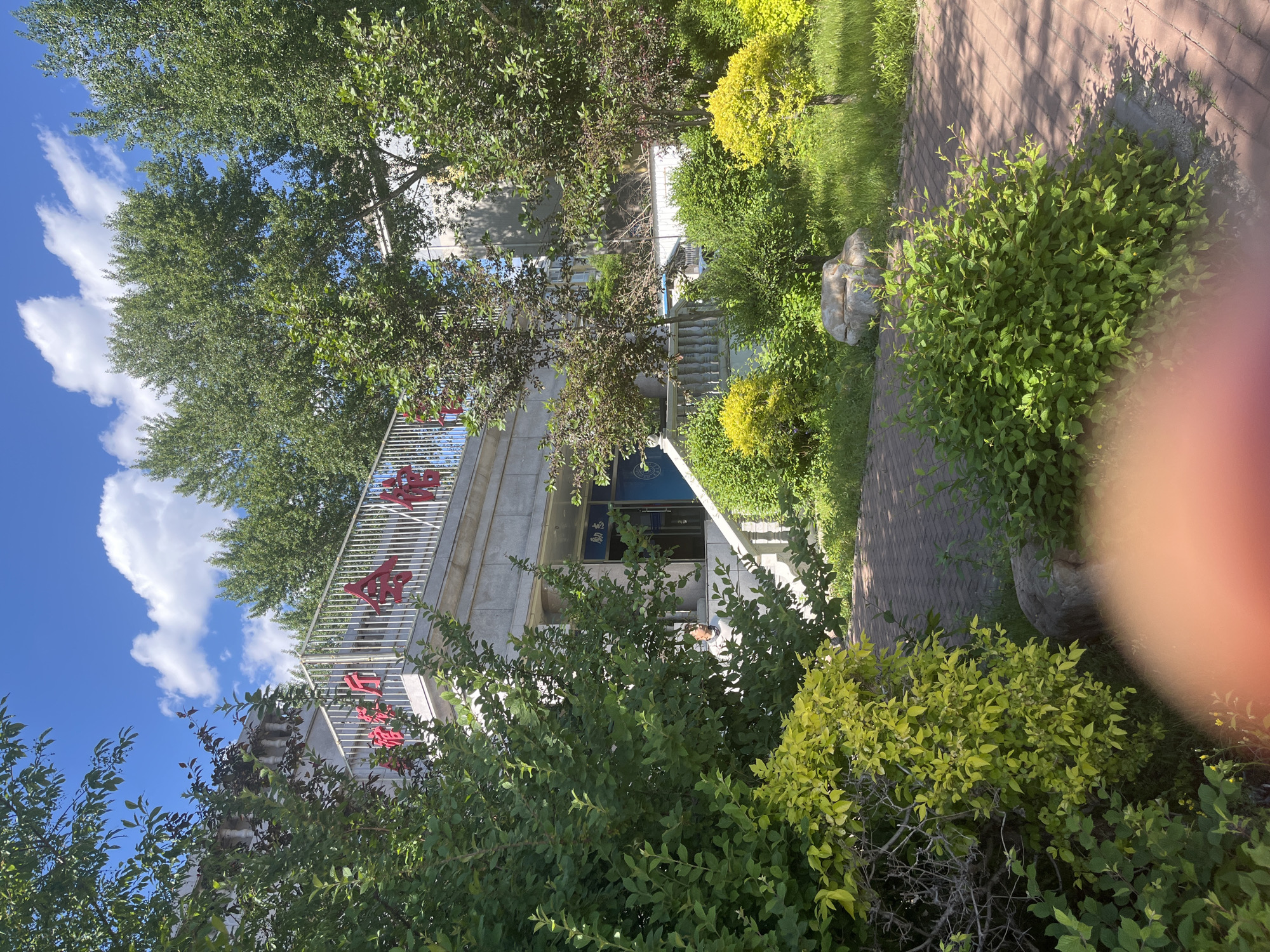 The image shows a stone building, likely a university or public institution, in Changchun City, China. The building has a balcony with a white metal railing and large red Chinese characters. It is surrounded by lush greenery, including trees, bushes, and manicured lawns. A brick pathway leads to the building's entrance. The sky is blue with scattered white clouds. Part of a finger is also in the picture's lower right, showing that the camera was close to the photographer.