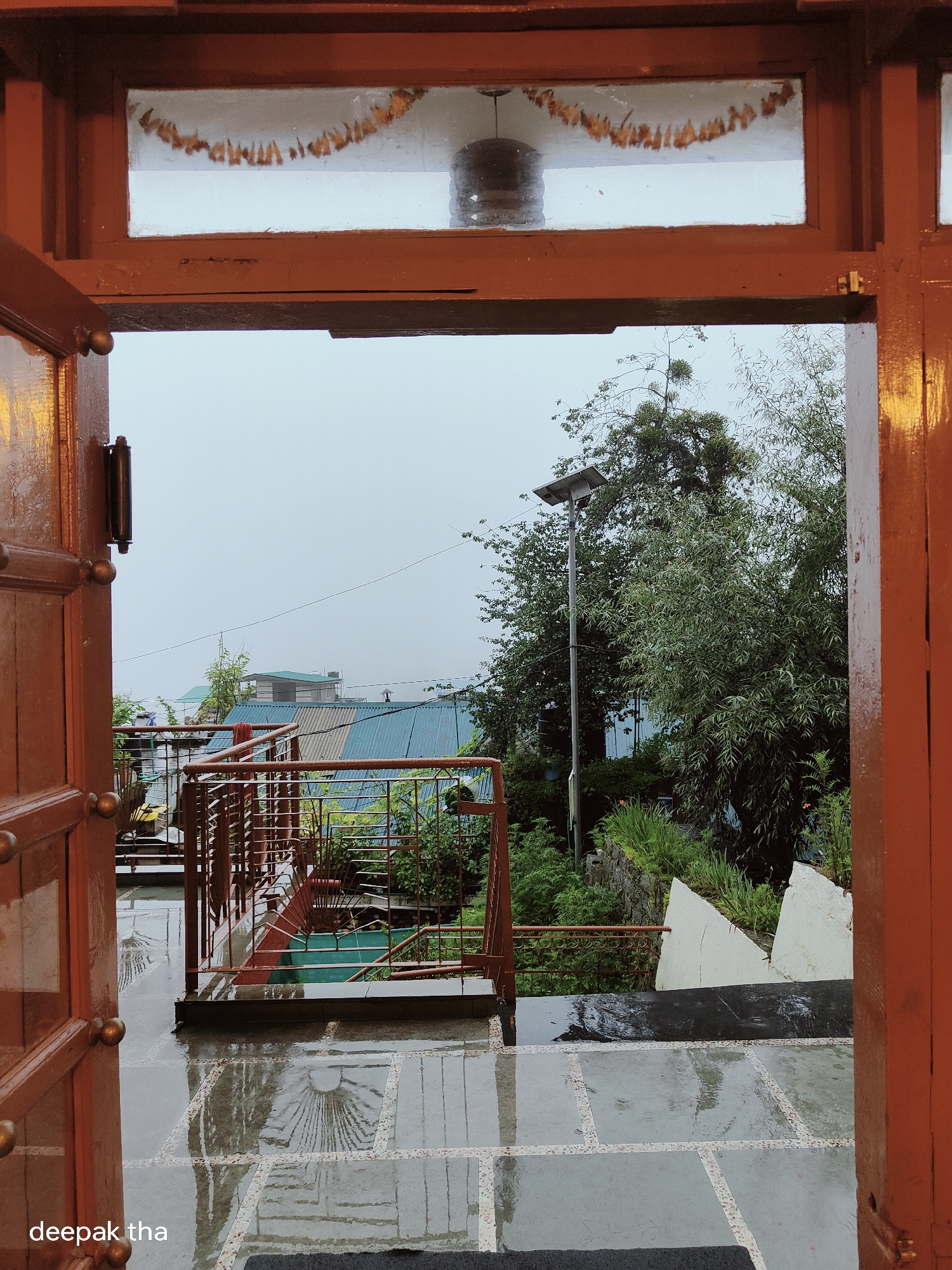 The image shows a view through an open wooden door frame, looking out onto a wet, tiled patio and a lush, green hillside. The sky is overcast. The patio is paved with large, square gray tiles with lighter borders. The tiles are wet and reflective, showing ripples from the rain. In the background, there is a reddish-brown metal railing leading down some steps, with some green plants visible below. Beyond the railing, rooftops of buildings can be seen, some with blue metal roofs. Trees and foliage are abundant, with a tall lamp post with a solar panel visible to the right of center. The overall impression is of a peaceful, slightly damp day in a mountainous area. The location is identified as Vashisht, India, a place known for its hot springs and scenic beauty.