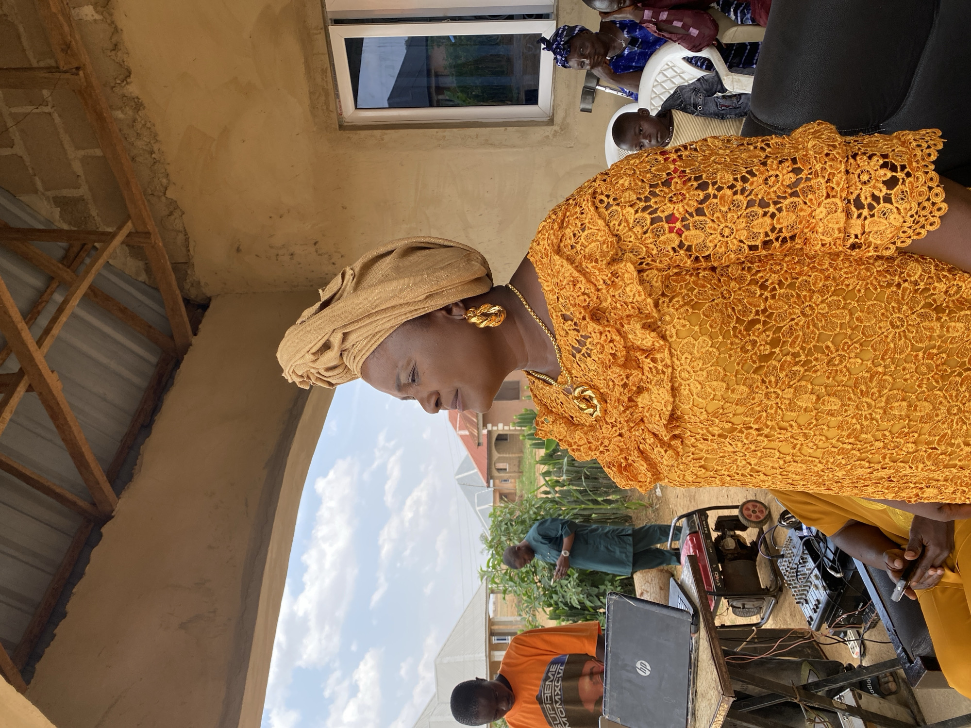 The image captures an outdoor setting in Barikin Sabon Gidan Kanat, Nigeria, depicting a gathering. The main subject is a woman in a gold-colored lace dress and headwrap, adorned with gold jewelry. She is positioned beside an equipment setup including a laptop, mixing console, and generator, which suggests a sound system setup. In the background, other individuals are visible, including seated people and a man in a green outfit. Buildings are seen farther in the background. The sky is partly cloudy.