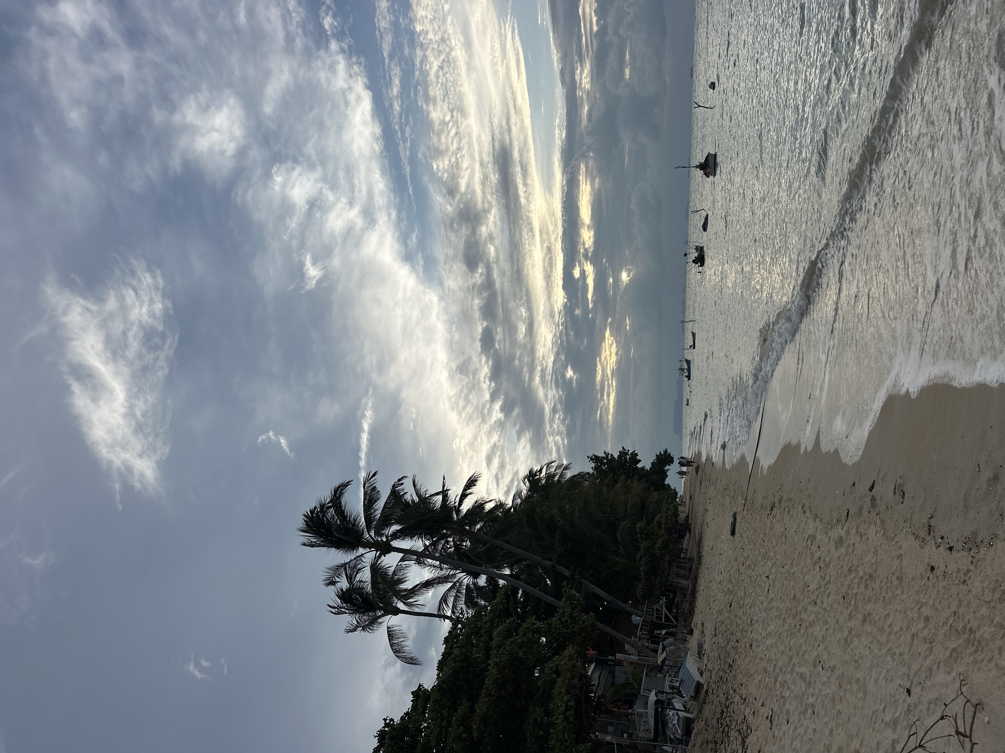 This image depicts a beach scene in the Ko Samui District of Thailand. The foreground shows a sandy beach with a few pieces of debris and some sparse vegetation. The ocean is to the right, with gentle waves lapping at the shore. Several small fishing boats are anchored in the water. In the background, there is a line of palm trees and other vegetation, with some buildings nestled amongst the trees. The sky is partly cloudy, with the sun peeking through the clouds, suggesting it is either sunrise or sunset. A few people are visible walking along the shoreline in the distance. The overall atmosphere is tranquil and serene.