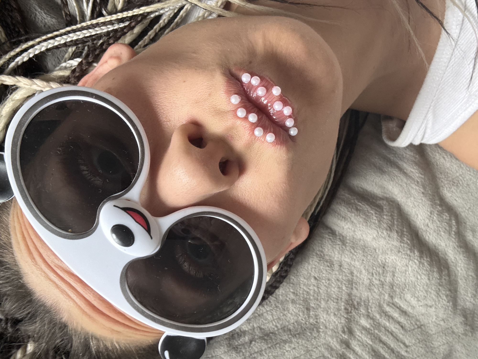 An overhead, slightly angled close-up portrait features a person lying down, showcasing their head and upper torso. The individual's face is adorned with unique accessories and makeup. They wear large, white, round sunglasses designed to resemble a panda's face, complete with black lenses, a small black nose, and a red mouth detail on the bridge, as well as small black ears on the outer frames. Behind the dark lenses, the eyes are obscured.
A striking feature is the lips, which are coated with a glossy substance and decorated with numerous small, white, pearl-like beads. These beads are meticulously arranged across both the upper and lower lips, creating a textured, avant-garde makeup effect. The person's hair consists of multiple thin, multi-toned braids or dreadlocks in shades of brown, blonde, and black, fanning out around their head.
A portion of a white t-shirt or top is visible on the right side of the frame, and the person rests on a soft, light gray or beige textured fabric, likely a blanket or duvet. The lighting appears to be bright and even, suggesting an indoor setting. The overall impression is one of artistic expression or a fashion statement. No text is visible in the image. This scene is set in Rusanovo, Russia.