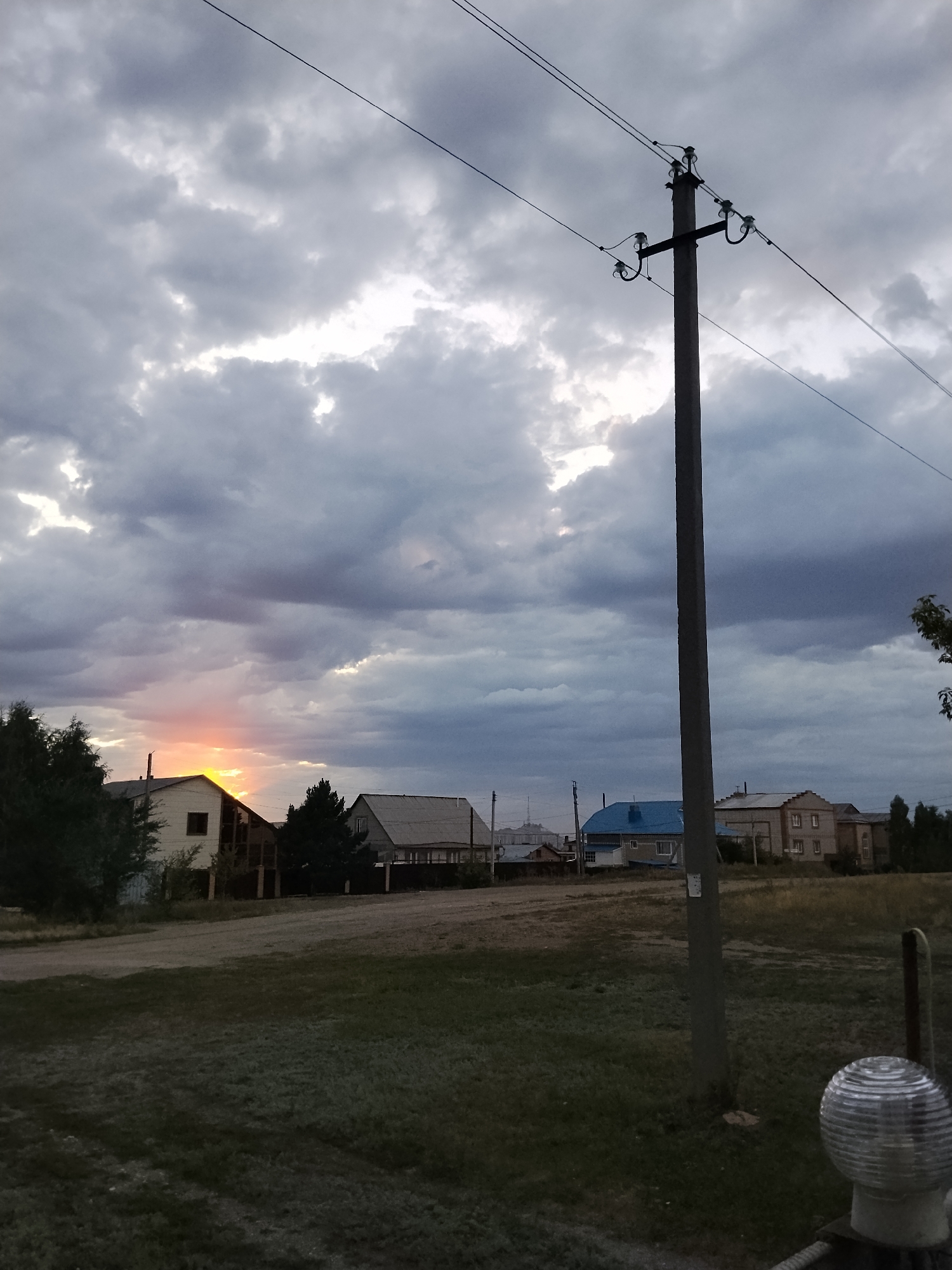 The image shows a rural scene with houses, a utility pole, and a dramatic cloudy sky at sunset. The sky is filled with dark, heavy clouds with patches of bright light breaking through, indicating the sun is setting or rising. A utility pole with wires stands prominently in the foreground, slightly off-center to the right. In the background, several houses of various sizes and architectural styles are visible, with a dirt road in front of them. Trees are scattered throughout the scene, some in front of the houses and some forming a darker line on the left side of the image. The ground in the foreground is covered in grass, with some bare patches. A circular, glass-like object with a ribbed surface is visible in the lower right corner of the image, likely part of a fence or a garden ornament. The overall impression is a quiet, overcast evening in a residential area. Given the context, this scene is likely from the city of Kokshetau, Kazakhstan, a region known for its rural settlements and natural landscapes.