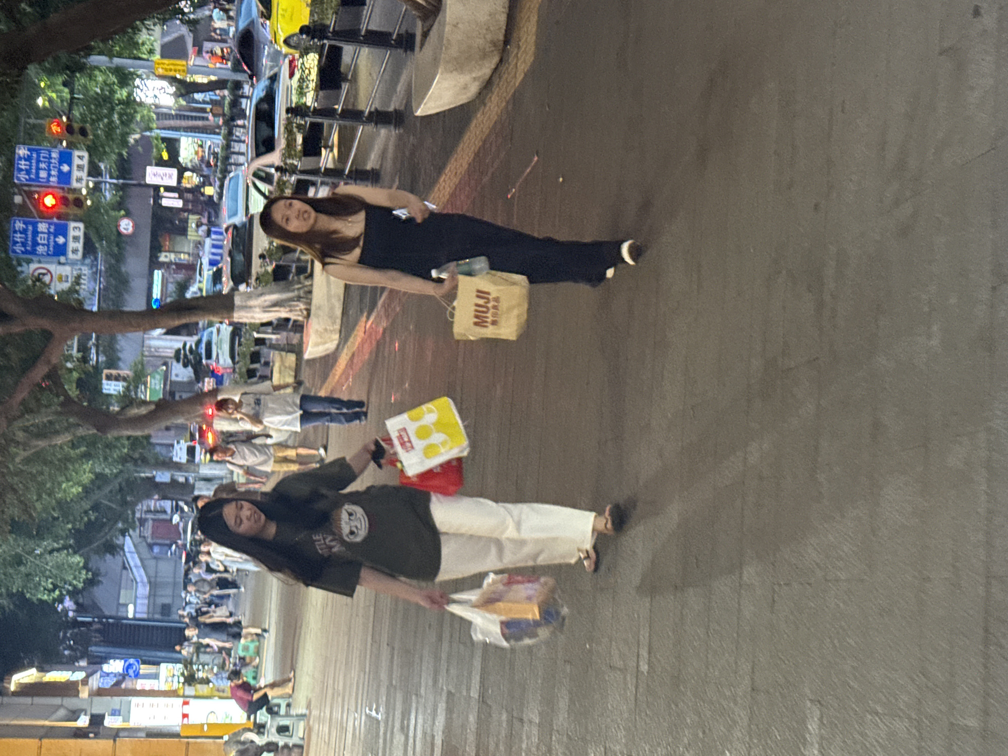 In Yuzhong District, China, two young women walk along a pedestrian street at dusk. The woman in the foreground on the left wears a dark green t-shirt with a graphic print and white pants. She carries a red shopping bag and a larger white shopping bag filled with items. The woman on the right wears a black spaghetti-strap maxi dress and carries a beige MUJI shopping bag and a bottle of water. Both women are walking away from the camera and appear to be engaged in shopping activities. The street is paved with cobblestones, and trees line the sidewalk. In the background, traffic lights are visible, and cars are driving on the road. Pedestrians and shops can be seen further down the street. The overall scene suggests a bustling urban environment during an evening stroll.