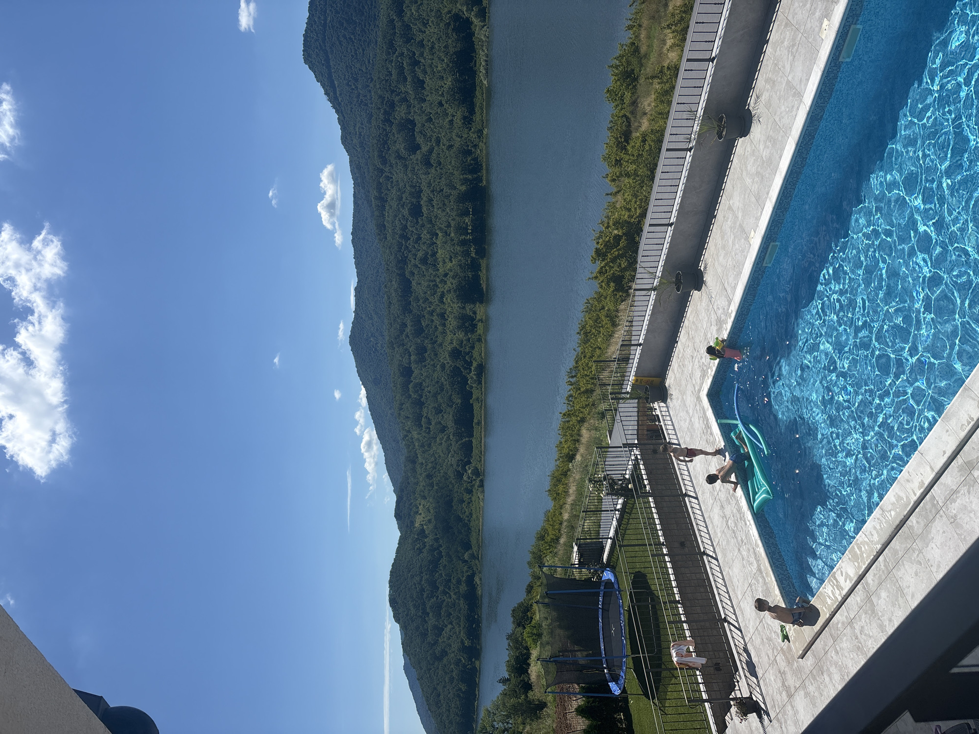 An outdoor, high-angle, full shot captures a serene summer day with a clear blue sky and fluffy white clouds. The scene is dominated by a vast, calm lake reflecting the bright sky, backed by lush, tree-covered mountains. In the foreground, a rectangular swimming pool with vibrant blue water sparkles under the sun. Several children are enjoying the water: two boys are in the pool, one on a green inflatable raft, and another is partially visible at the edge. Another child stands at the edge of the pool, looking towards the water. A trampoline with a blue net is visible on the green lawn next to the pool, and a towel hangs on a fence. The pool area is paved with light-colored tiles and enclosed by a dark metal fence. The environment suggests a residential property with a stunning natural backdrop.