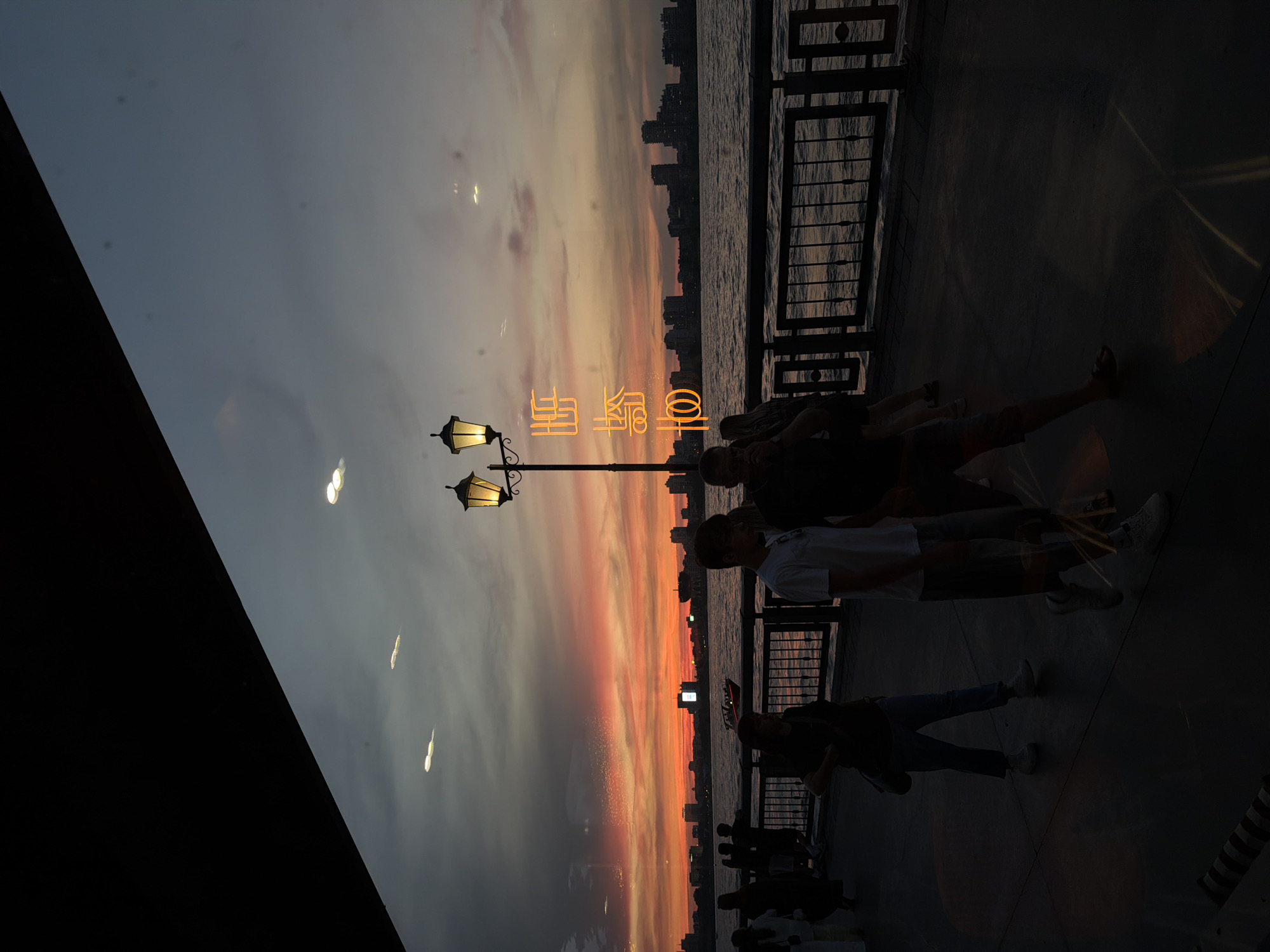 This is a vertical, medium-shot, eye-level photograph taken at dusk along a waterfront promenade, possibly in Kazan, Russia, given the context. The scene depicts a vibrant sunset with fiery orange and pink hues streaking across a twilight sky, casting a warm glow over the water and silhouetting a city skyline in the distance.
In the foreground, a dark, angled structure, possibly a building overhang or awning, occupies the top left portion of the frame, creating a strong diagonal line that divides the sky from the lower part of the image. A lamppost with two illuminated, ornate lamps stands prominently on the right side of the walkway, casting a warm light. To its left, a large, bright orange neon sign with stylized Chinese characters is visible, appearing to be mounted on the lamppost or an adjacent structure.
A promenade or walkway runs along the waterfront, with a black metal railing separating it from the dark, rippling water. Several people are walking along the promenade, mostly appearing as silhouettes due to the low light and backlight from the sunset. In the middle ground, two men are walking side-by-side, one wearing a white t-shirt and the other a darker top. Further back, other individuals and groups are faintly discernible, some standing and others walking. A small boat with a red light is visible on the water.
The ground of the promenade is dark, with scattered patches of light reflecting on it, possibly from the neon sign and the lamppost. The overall atmosphere is one of a tranquil evening by the water, with the natural beauty of the sunset enhanced by the artificial light of the lamps and the neon sign.