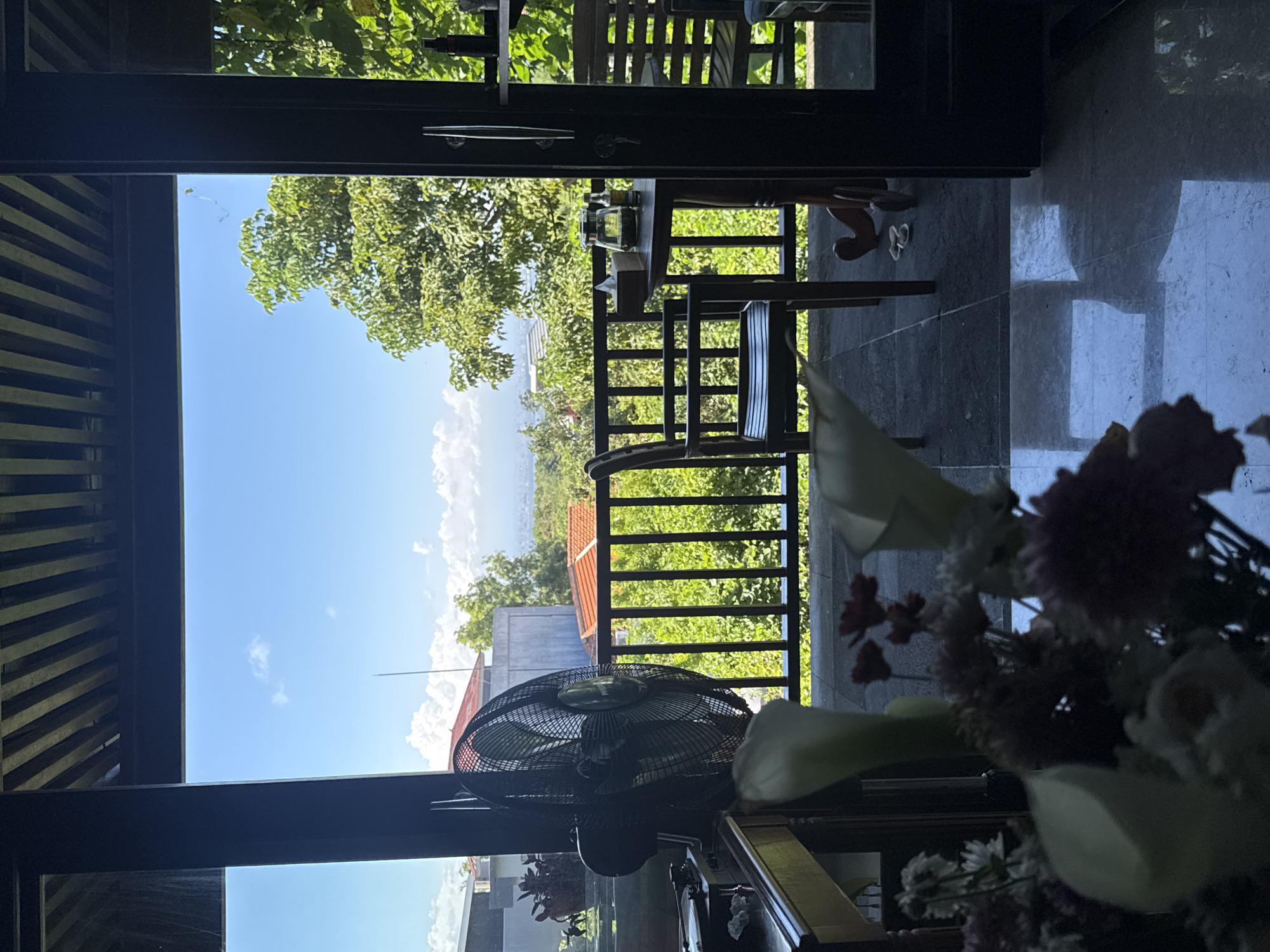 The image shows a view from inside a building in Kuta Selatan, Indonesia, looking out onto a terrace. The terrace has a dark wood railing and furniture. A black electric fan is visible indoors near the window. The outdoor scenery features lush green trees, and buildings. The sky is blue with white clouds. Indoors there is an arrangement of flowers in the foreground. The building structure and window frame are made of dark material, possibly wood or metal.