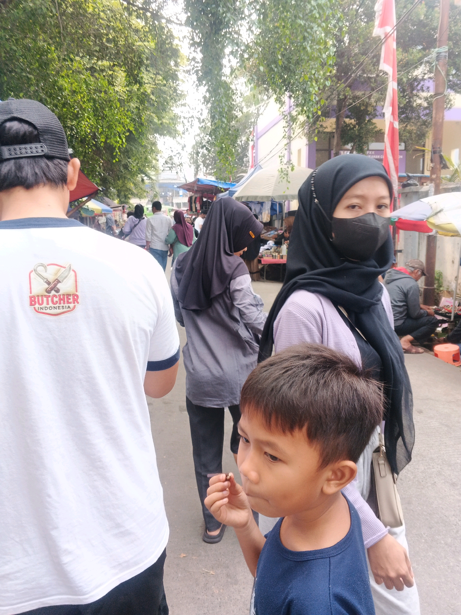 The image captures a street scene in Curug Kulon, Indonesia, with a focus on several individuals and the surrounding market-like environment. In the foreground, a young boy in a blue t-shirt is seen from the chest up, looking to his left with a piece of food in his right hand, which he is bringing to his mouth. Behind him and to his right, a woman wearing a black hijab and a black face mask is visible from the chest up. She is wearing a light purple cardigan over a black garment and has a cream-colored bag slung over her shoulder. To the left of the boy, the back of a person wearing a white t-shirt with a "BUTCHER INDONESIA" logo and a black baseball cap is visible. Further in the background, other individuals are walking and interacting, with market stalls and awnings lining the street. Trees with green foliage are prominent in the upper portion of the image, suggesting an outdoor, possibly warm, environment. The lighting indicates it is daytime.