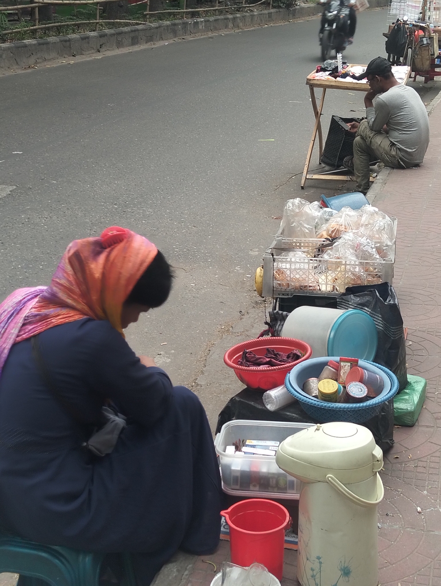 In Dhaka, Bangladesh, a woman with an orange and purple headscarf sits on a green plastic stool next to a variety of goods laid out for sale on the sidewalk. These include bagged foods in a wire basket, containers, bowls, and a large thermos. A man sits further down the sidewalk next to a folding table displaying items, possibly clothing. A motorbike passes on the adjacent road.