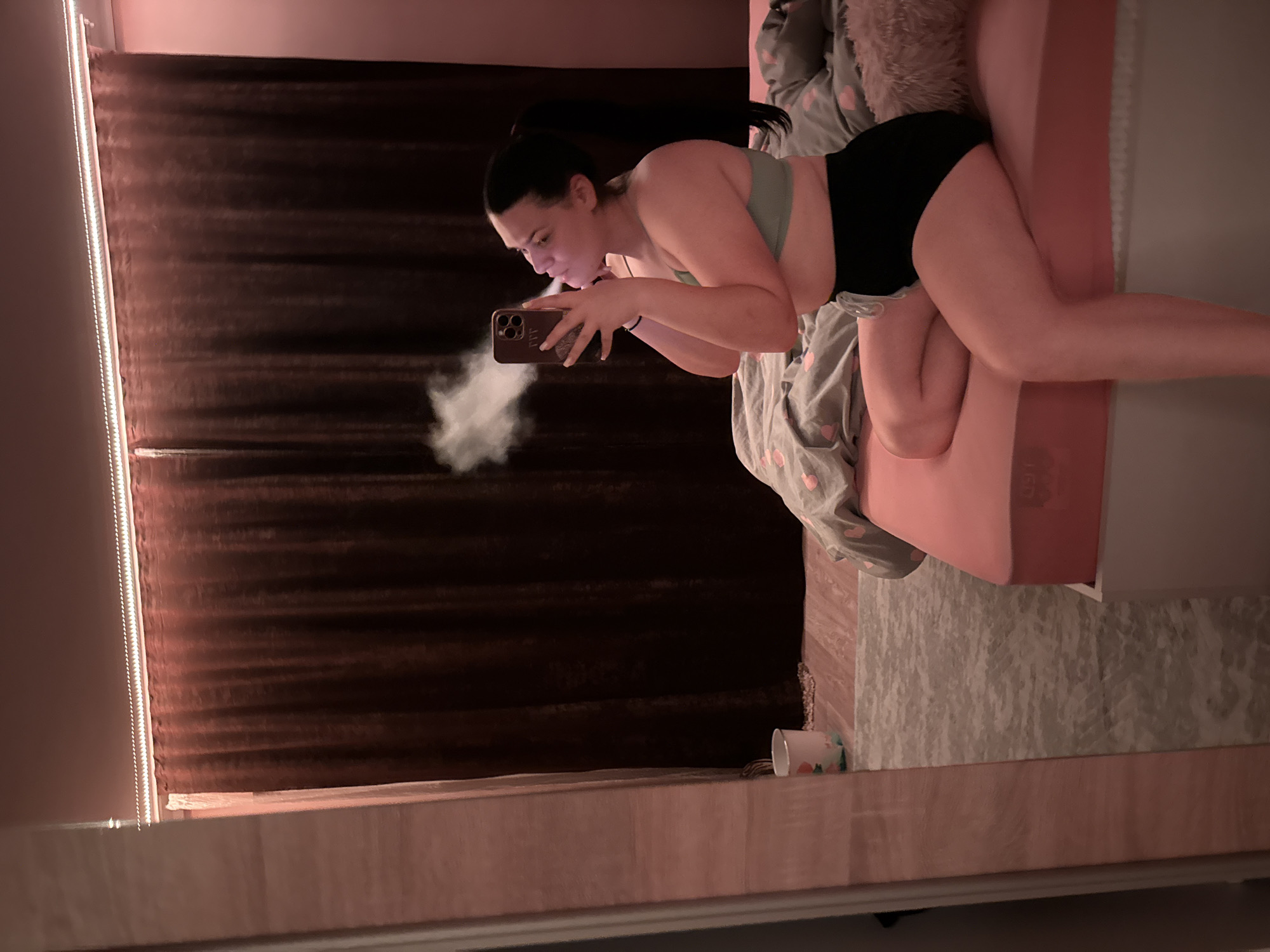 In a bedroom, likely located in Rogosh, Bulgaria, a young woman with dark hair pulled into a ponytail sits on the edge of a bed. She is wearing a light green sports bra and black shorts. The woman is holding a smartphone in her right hand, angled towards her face, and appears to be taking a selfie or recording a video, as a cloud of white vapor is visible emanating from her mouth and obscuring part of the phone. Her left hand is raised, possibly to adjust her hair or the phone. The bed is covered with a pink sheet and grey bedding with a heart pattern. Behind the woman, dark brown velvet curtains hang, and a string of white LED lights is mounted on the ceiling above them, casting a warm glow. To the left of the frame, a large mirror reflects the scene, and part of a pink and white patterned trash can is visible on the floor in the reflection. The overall lighting in the room is dim and atmospheric, with a pinkish hue dominating the walls and light sources.