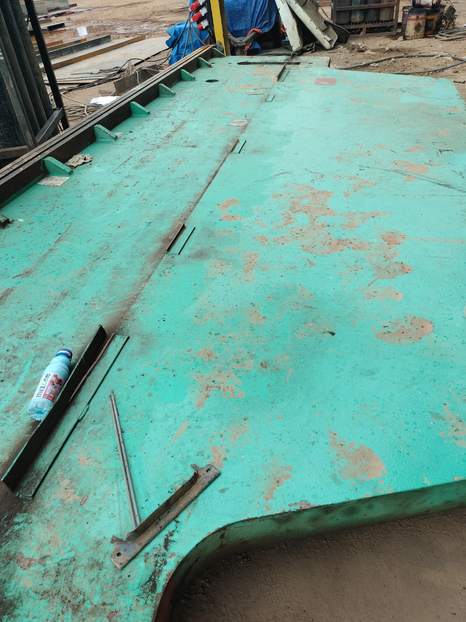 An outdoor scene in Batam, Indonesia, displays a large, flat, turquoise-painted metal surface, likely part of a construction or industrial site. The surface is weathered, with areas of rust and dirt accumulation, suggesting outdoor exposure. Several metal beams and structural elements are visible along the left edge of the frame, and to the rear, various construction materials, including tarps, pipes, and containers, are present. A partially filled bottle of "Mizone" brand flavored water rests on the metal surface. Two thin metal rods and a rusted bracket are also present on the surface. The ground surrounding the metal platform appears to be dirt and gravel, indicative of an industrial or construction environment.