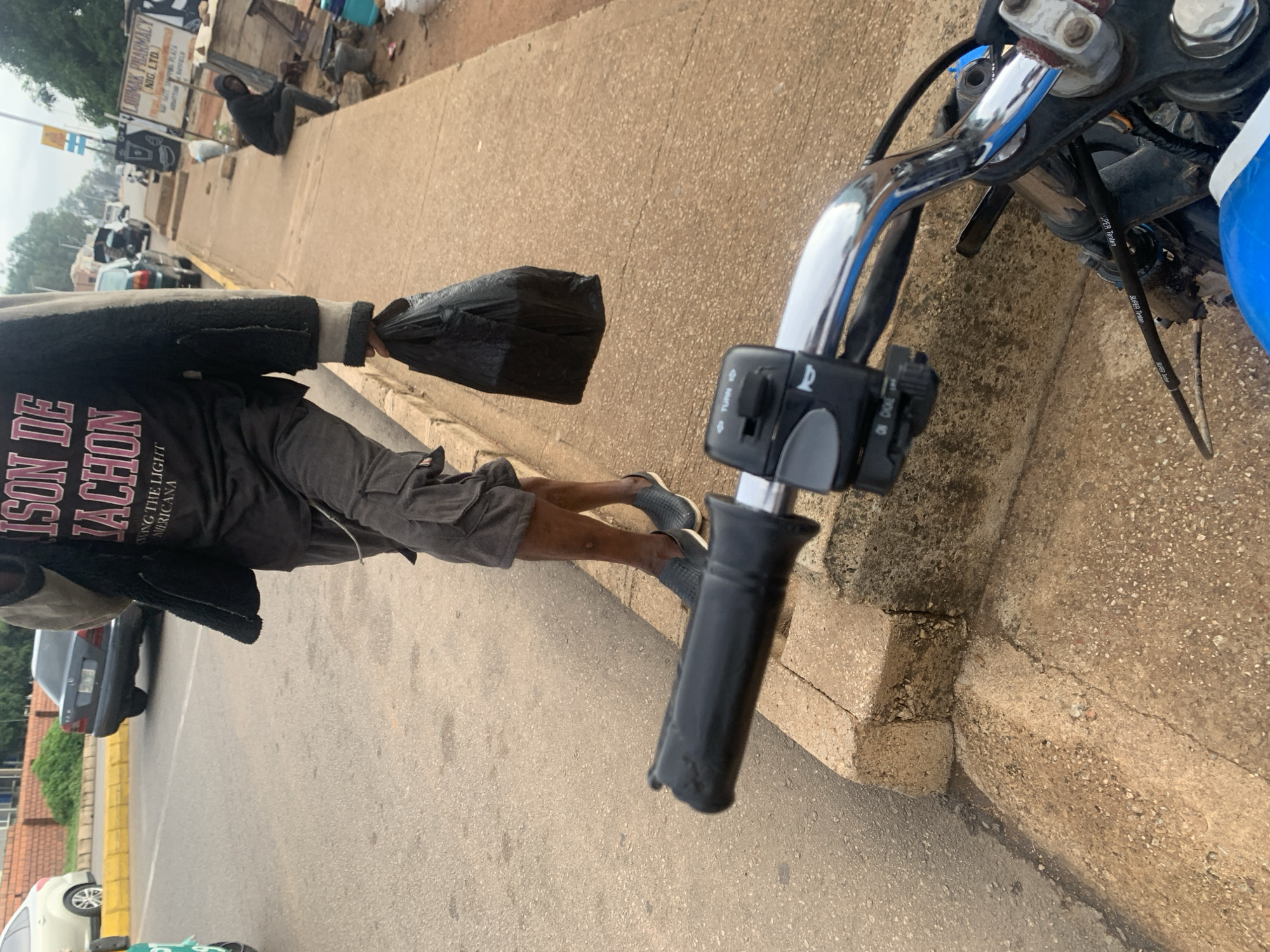 This image captures a daytime street scene in Barakin Lerawi, Nigeria, from the perspective of someone on a motorcycle. In the foreground, chrome motorcycle handlebars with black grips and control switches are prominent. The switches are labeled "TURN," "ON/OFF" (or "ON DIOKE" as per OCR), and a 'P' symbol. A cable marked "SUPER Tenten" is also visible, running alongside the handlebars.
A person, likely the motorcyclist, is centrally featured, wearing a dark t-shirt with red and white text ("ISON DE JACHON," "WING THE LIGHT," "MERICANA"), dark cargo shorts, and a dark, textured jacket. They are holding a black plastic bag in their left hand. Their feet, clad in dark shoes, are visible near the concrete curb.
The street is paved, with a concrete sidewalk and curb running along the right side of the frame. In the midground, cars are parked and moving, including a dark sedan and a white vehicle. Further down the street, another person is sitting on the ground next to a structure, with a small animal (possibly a dog) and a blue container nearby. Various signs are visible in the background, including one for "LAMAK PHARMACY NIG. LTD." and another with "ART TURBANFELD." The lighting suggests an overcast or partly cloudy daytime, with a generally bright but diffused light. The scene depicts a common street environment.
