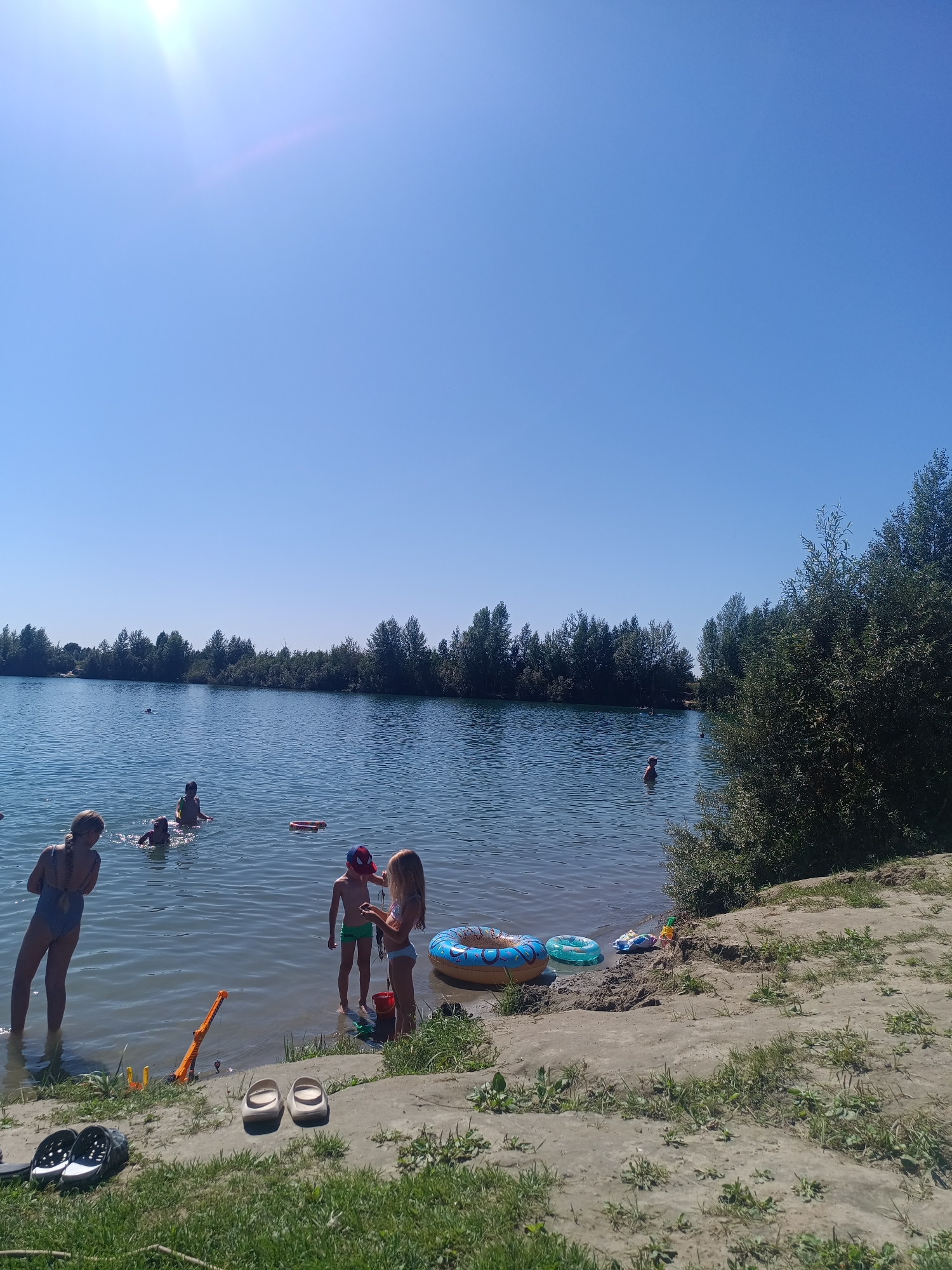 In Mochishche, Russia, on a clear, sunny day, a group of people are enjoying leisure activities at a lake. The foreground features a sandy embankment with green grass and several pairs of shoes scattered on the ground. Among these are black and white slide-style sandals and a pair of beige slides. A bright orange toy crane and some yellow plastic toys are also visible near the water's edge, along with a red bucket. Further out in the shallow water, two children, a boy wearing green shorts and a girl with blonde hair wearing a bikini, are standing and interacting. The boy is holding a string, possibly engaged in play. To the right of them, a blue inflatable ring with an orange pattern floats near the shore. Another smaller, green inflatable ring is also present in the water. Further into the lake, several other individuals are visible swimming or wading, including a person in a red cap. The lake stretches to the horizon, bordered by a dense line of green trees. The sky is a clear, bright blue, with the sun creating a lens flare in the upper left quadrant of the image.
