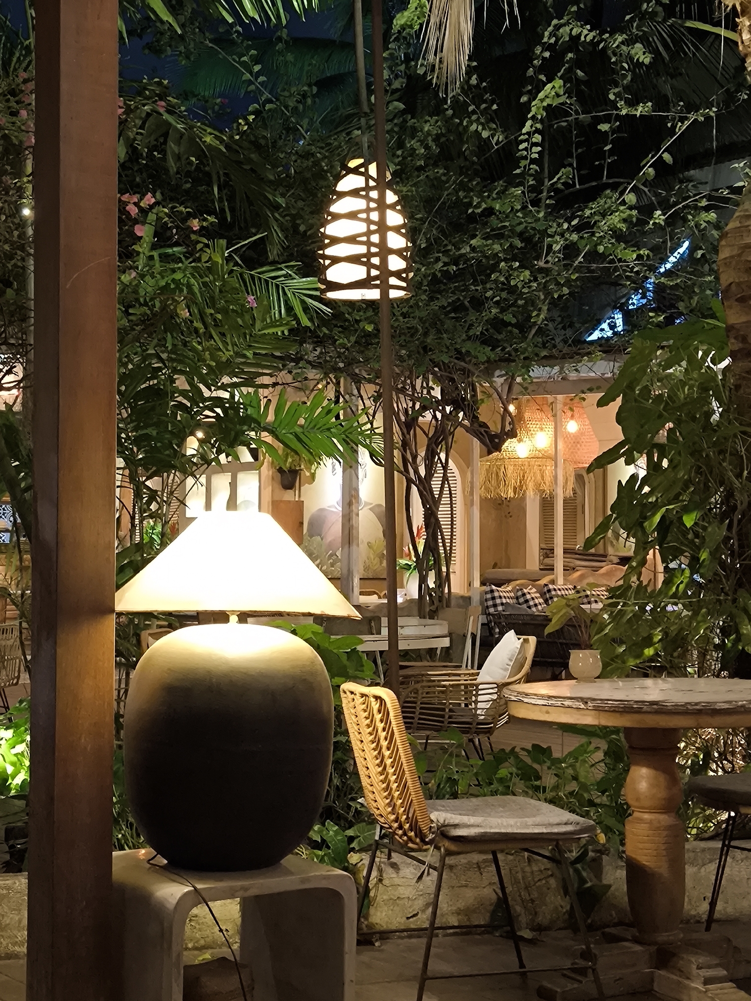 The image captures a cozy and verdant outdoor or semi-outdoor dining/lounge area at night, likely a restaurant or cafe in North Kuta, Indonesia. The setting is lush with dense tropical foliage and various plants, creating a serene, jungle-like ambiance.
In the immediate foreground on the left, a large, dark-colored table lamp with a wide, glowing conical shade sits on a light-toned, rectangular plinth. Adjacent to it is a substantial wooden post. Moving into the mid-ground, a woven wicker chair with a light cushion is positioned next to a rustic, round wooden table with a sturdy pedestal base. A small white ceramic pot with a plant rests on the table. Another decorative lamp, a vertical fixture with a woven, lantern-like shade, illuminates the central area. The surrounding space is rich with green leaves, vines, and some delicate pink blossoms.
In the background, a building with arched openings, some fitted with wooden shutters, is visible. Inside, warm-toned pendant lights, crafted from materials resembling straw or wicker, cast a soft glow. Seating areas with checkered cushions are discernible within the interior space. The overall aesthetic is bohemian and tropical, enhanced by the abundant greenery and natural material furnishings. No people are visible, and no specific activities are taking place, but the setup is clearly for dining, relaxation, or socializing. The dark sky confirms it is nighttime, with all illumination coming from the various lamps and interior lighting, contributing to a warm, inviting, and tranquil atmosphere. There is no visible text.