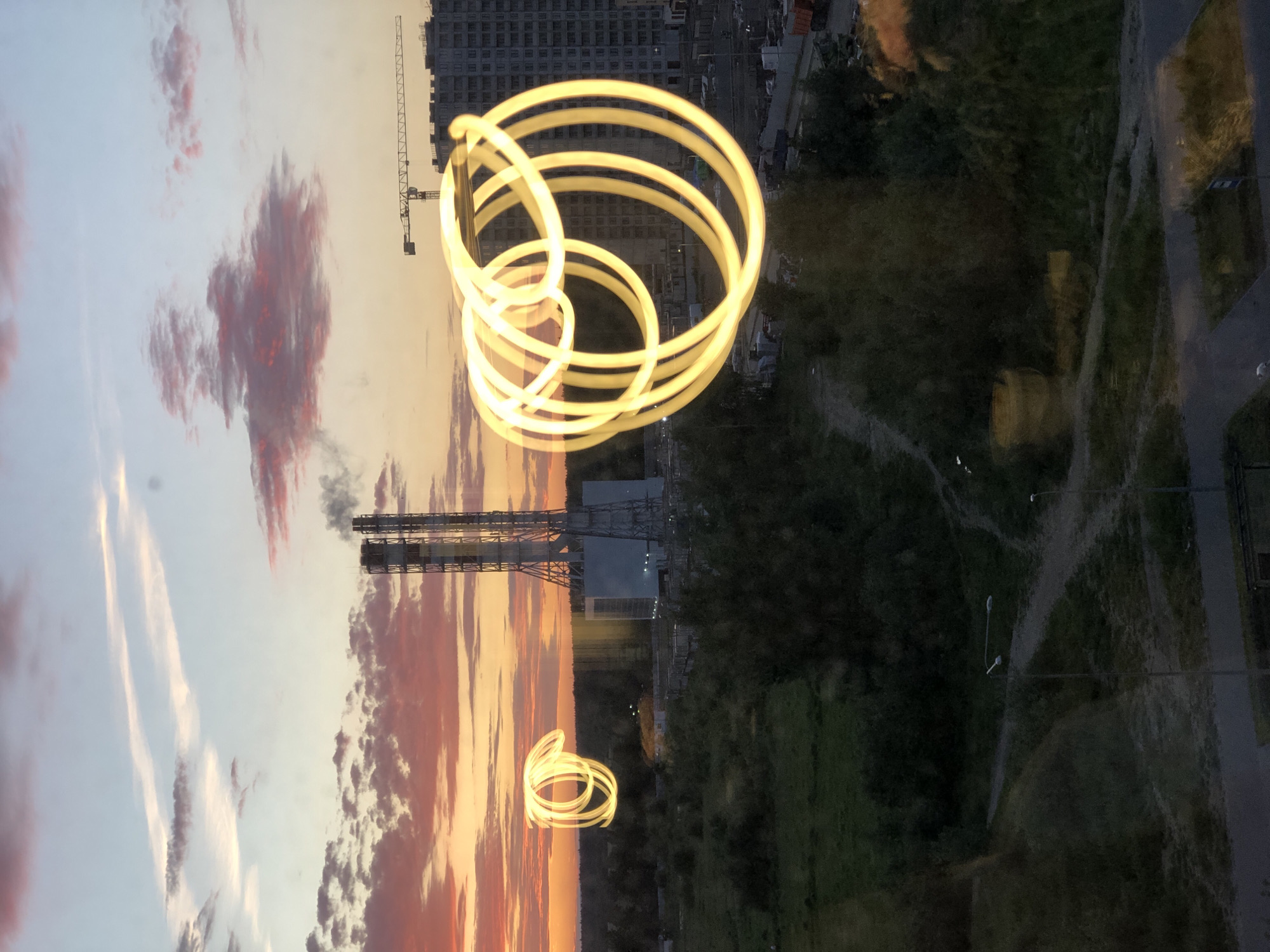 The image depicts an outdoor scene at dusk with a sky painted in hues of orange, yellow, and pink from the setting sun. Two tall, industrial chimneys with smoke billowing from them are visible in the center-left of the frame, suggesting the presence of a power plant or factory. In front of these chimneys, a circular light painting of glowing yellow lines creates an abstract, swirling effect. To the right of the chimneys, a tall building under construction, possibly an apartment complex, is visible with a crane positioned above it. The foreground shows a landscape of green grass and trees, with a road and pathways winding through it. The overall impression is a blend of industrial infrastructure, ongoing construction, and a natural landscape illuminated by the soft light of twilight and an artistic light display. The location is the area near the **KPP-2 power plant in the Sosnovy Bor district of Saint Petersburg, Russia**. There are no identifiable people in the image.