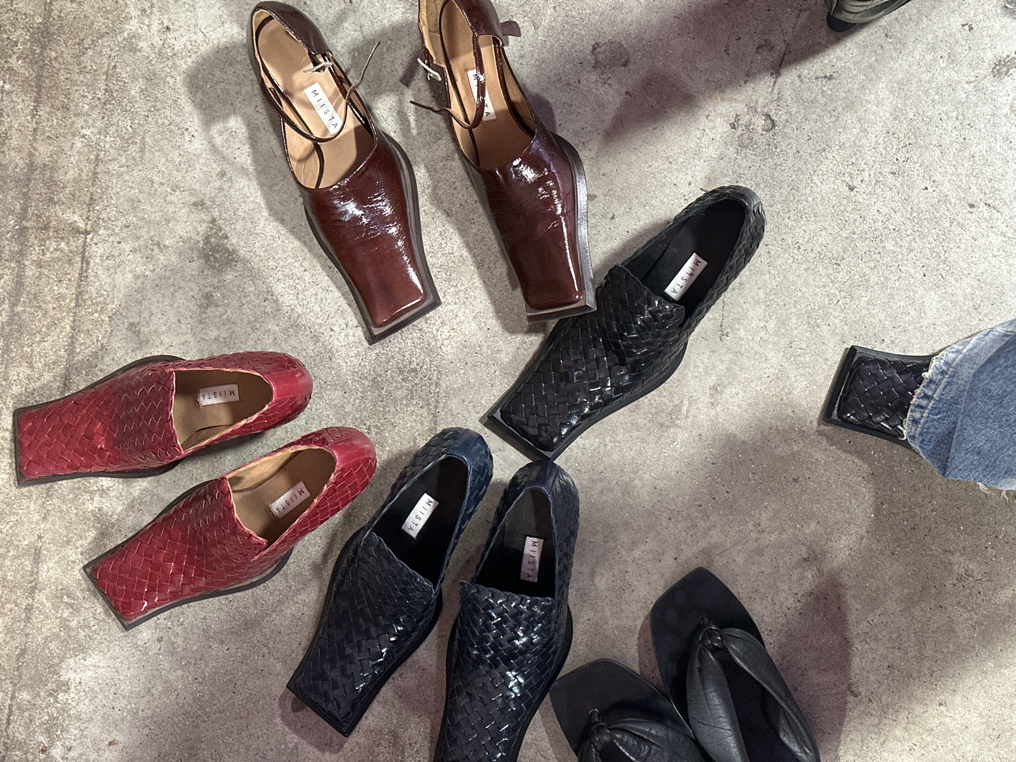 The image depicts an assortment of women's shoes arranged on a concrete surface, likely an indoor floor. Two pairs of burgundy-colored patent leather heeled shoes with ankle straps are positioned at the top. Adjacent to them are two pairs of black woven-leather pointed-toe loafers. To the left, two pairs of red woven-leather loafers are visible. Near the bottom, two pairs of black flat thong-style sandals. A leg clad in blue jeans is visible, with one shoe. The labels of the shoes are marked "MIISTA". Given the label and product focus, the setting is likely a studio or showroom showcasing shoe designs, but there is no indication that this is in London. The environment is uncluttered, directing attention to the footwear on display.