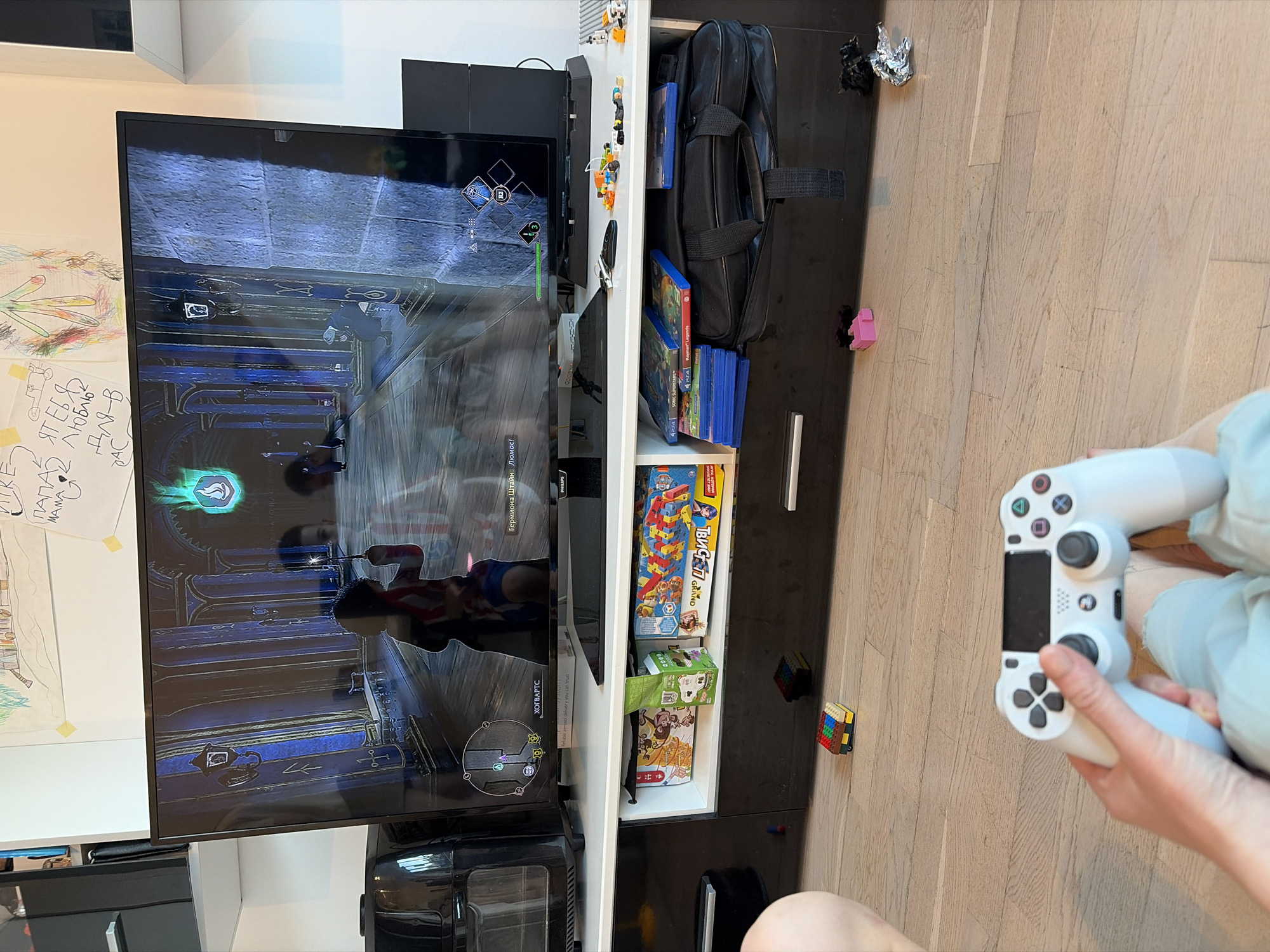 The image shows a person playing a video game on a television. The game displayed on the screen appears to be set in a dark, ornate interior with blue hues. The person is holding a white PlayStation 4 controller.
To the right of the television is a white shelf unit filled with video games, a black laptop bag, and board games. A few LEGO blocks are scattered on the wooden floor. A whiteboard with children's drawings and written notes is visible to the left of the television.