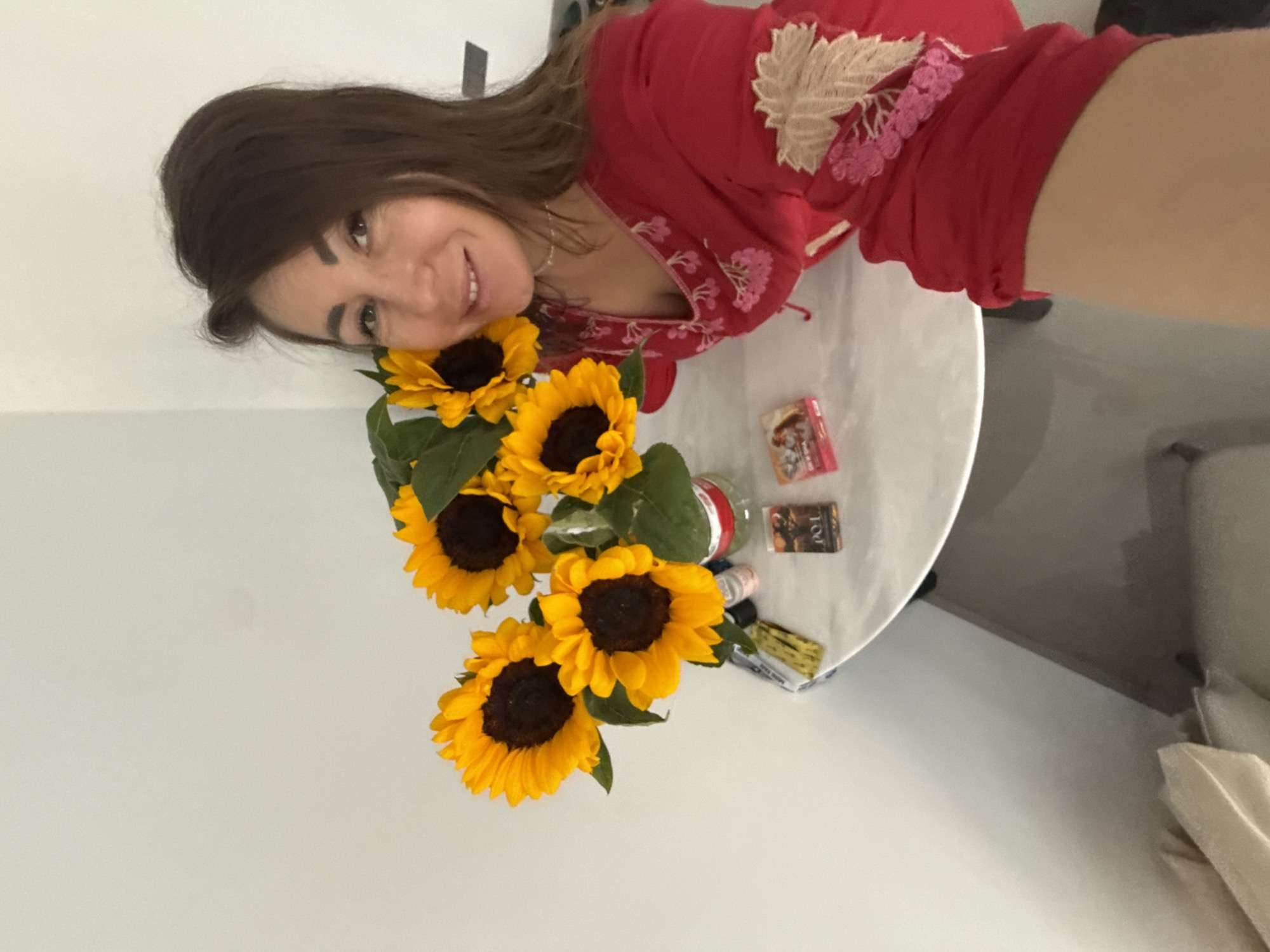 A woman with dark hair is taking a selfie in a room with white walls. She is wearing a red shirt with floral embroidery. In front of her is a round white table with a bouquet of sunflowers in a glass vase. Several small items are on the table, including what appear to be packets of cards or candy. The background is plain and uncluttered.