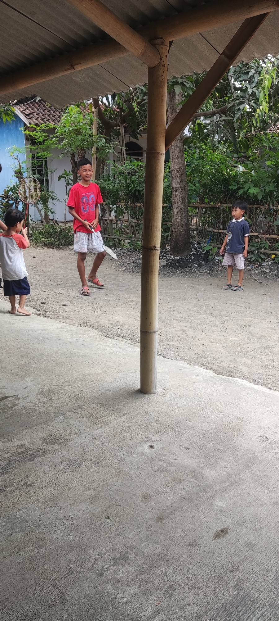 A low-angle, eye-level shot captures three young boys in Karangwangun, Indonesia, engaged in a game of badminton in a dusty, outdoor area. The scene is framed by a rustic bamboo structure with a corrugated roof overhead.

On the left, a boy wearing a red and white t-shirt and dark shorts stands barefoot, facing away from the camera and towards the other boys. He holds a badminton racket in his right hand.

In the center, a boy in a red t-shirt with "86" printed on it and patterned shorts stands, also barefoot, holding a badminton racket in his right hand. He appears to be smiling and looking towards the boy on the left.

On the right, a boy in a dark blue polo shirt and light-colored shorts stands facing left, holding a badminton racket in his right hand. He is looking towards the other boys. He wears dark sandals.

The ground is a mixture of concrete and packed dirt. Green foliage and trees are visible in the background, along with a bamboo fence and a blue-painted building. The overall atmosphere suggests a casual, outdoor recreational activity in a residential or communal outdoor space.