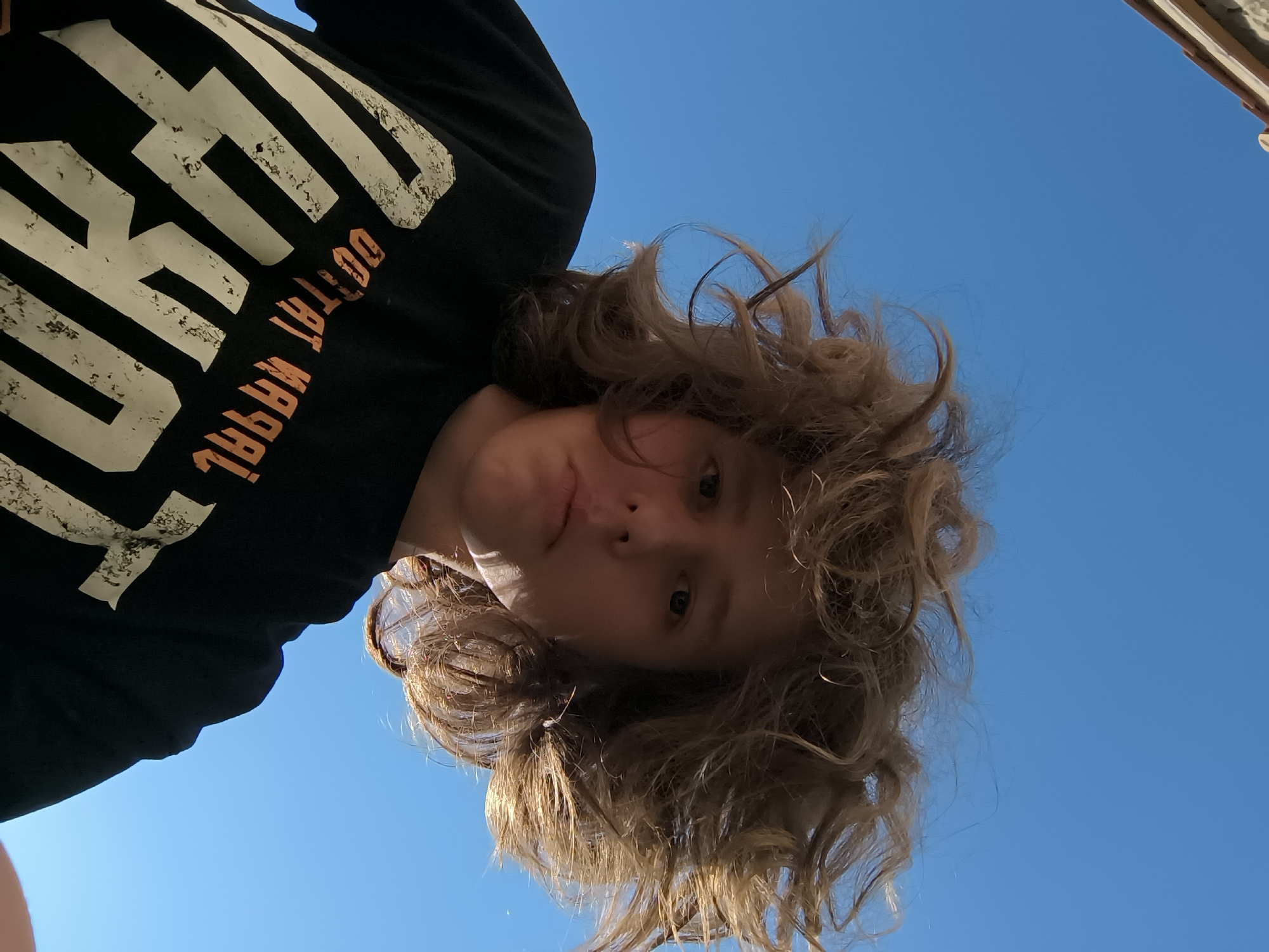 A person with curly brown hair and fair skin is looking down at the camera, wearing a black t-shirt with white lettering and orange text reading "DO TAT WRRAL." The background is a bright, clear blue sky. Part of a window or structure with a beige-colored exterior is visible in the upper right corner of the frame. The image is taken from a low angle, making the person appear to be towering over the viewer. The exact location in Pélussin, France, cannot be determined from the image alone, as there are no recognizable landmarks.