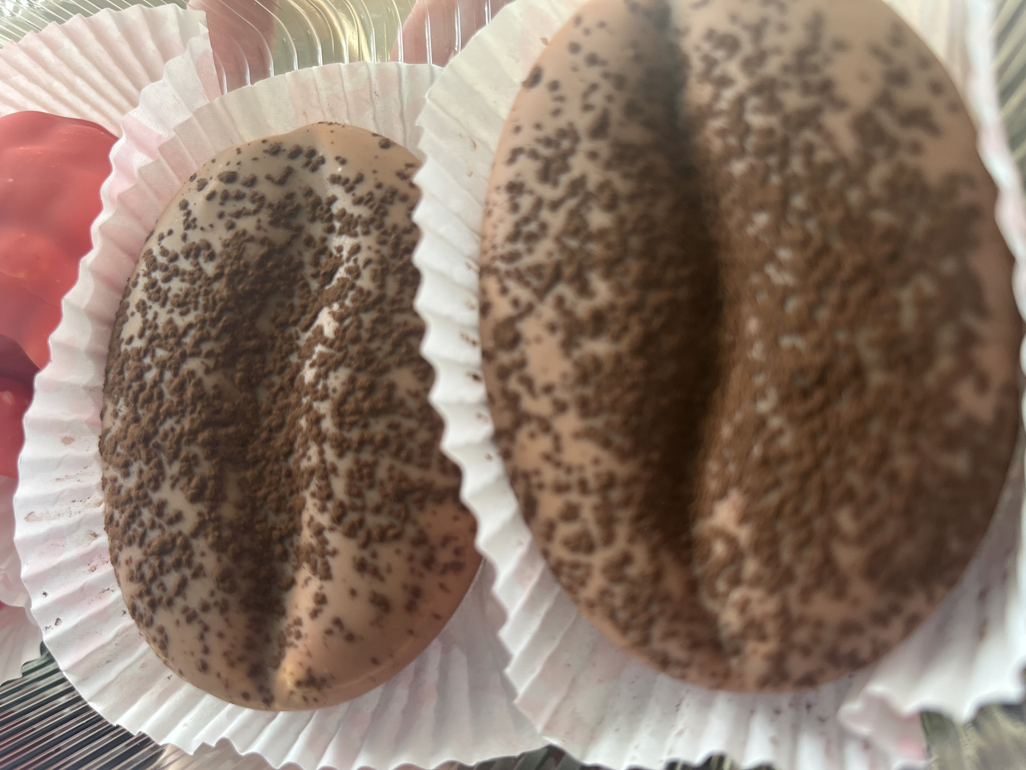 The image shows two chocolate cookies shaped like coffee beans, each resting in a white fluted paper liner. The cookies are a light brown color and are sprinkled with a darker brown powder, resembling cocoa or coffee grounds. A faint crease runs down the center of each cookie, enhancing the coffee bean shape. In the upper portion of the image, a blurry red object is visible behind the top cookie. The cookies are placed in a clear container, suggesting they are for sale or display. There are no people visible in the image. The scene appears to be indoors, possibly in a bakery or confectionery shop. The location in Desyatikhatka, Russia is not visually identifiable from the provided image.
