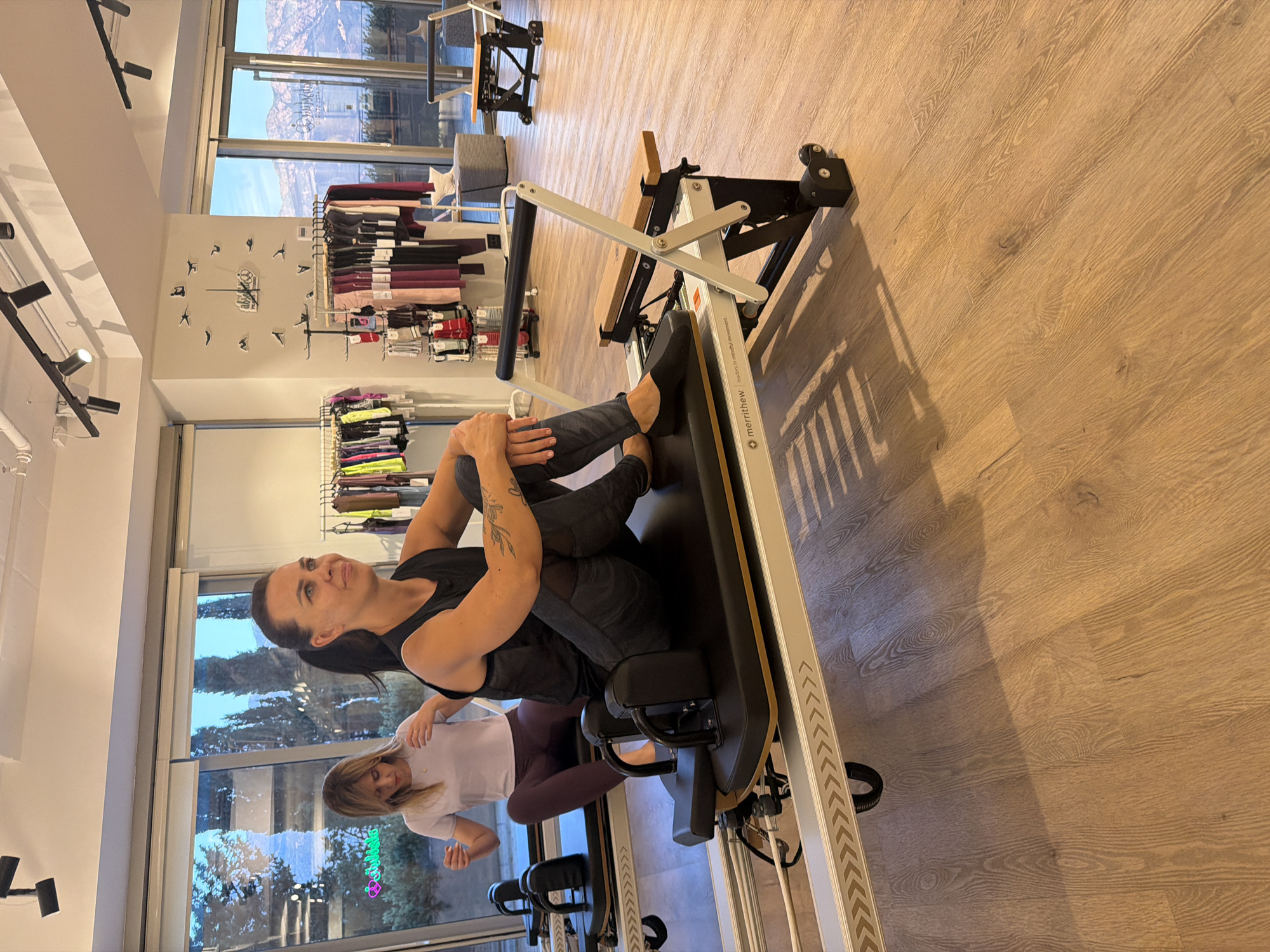 The image depicts two women engaged in a Pilates session in a studio located in Boreti, Montenegro. The studio has large windows overlooking a scenic view of mountains and water. In the foreground, a woman with dark hair is seated on a Pilates reformer machine, wearing a black tank top and patterned leggings. She has tattoos on her left arm and is looking to her right with a slight smile. Another woman with blonde hair is seated on a second Pilates reformer machine behind her, wearing a white t-shirt and purple leggings. She is looking forward and her hands are resting on the machine. The studio is well-lit and features a modern interior with wooden flooring and racks of athletic wear displayed on the wall. The Pilates reformers are professional-grade equipment, with one in the foreground featuring the "Merrithew" logo, indicating a specific brand. The overall atmosphere suggests a fitness or wellness center.