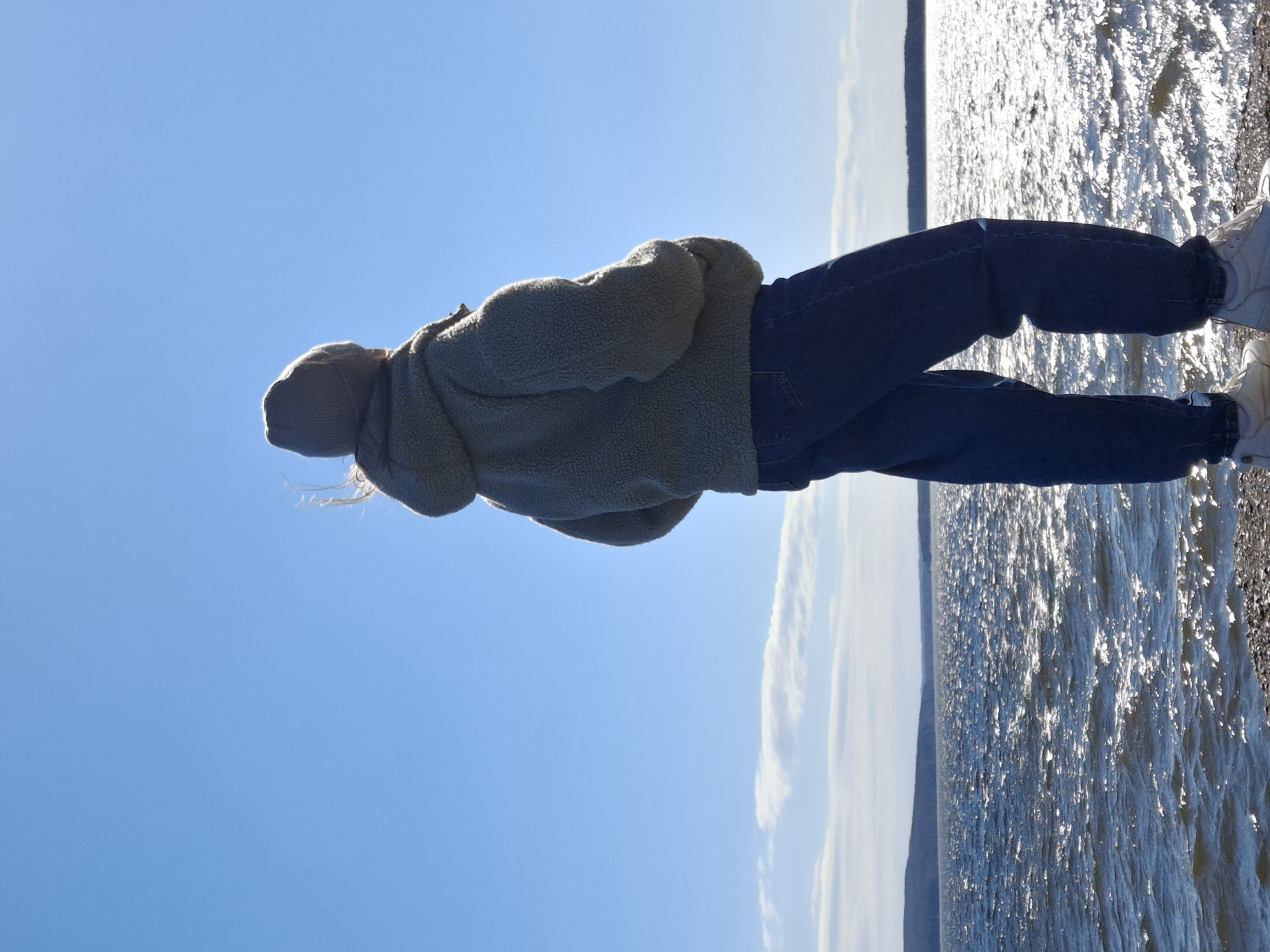 A person is standing next to a body of water, likely a river or lake, on a sunny day. They are wearing jeans, a jacket and sneakers. The background includes the water with bright sunlight reflecting off it, and a clear blue sky. The person's back is to the viewer, and they appear to be looking out at the water. The location is likely in Chaikovsky, Russia.