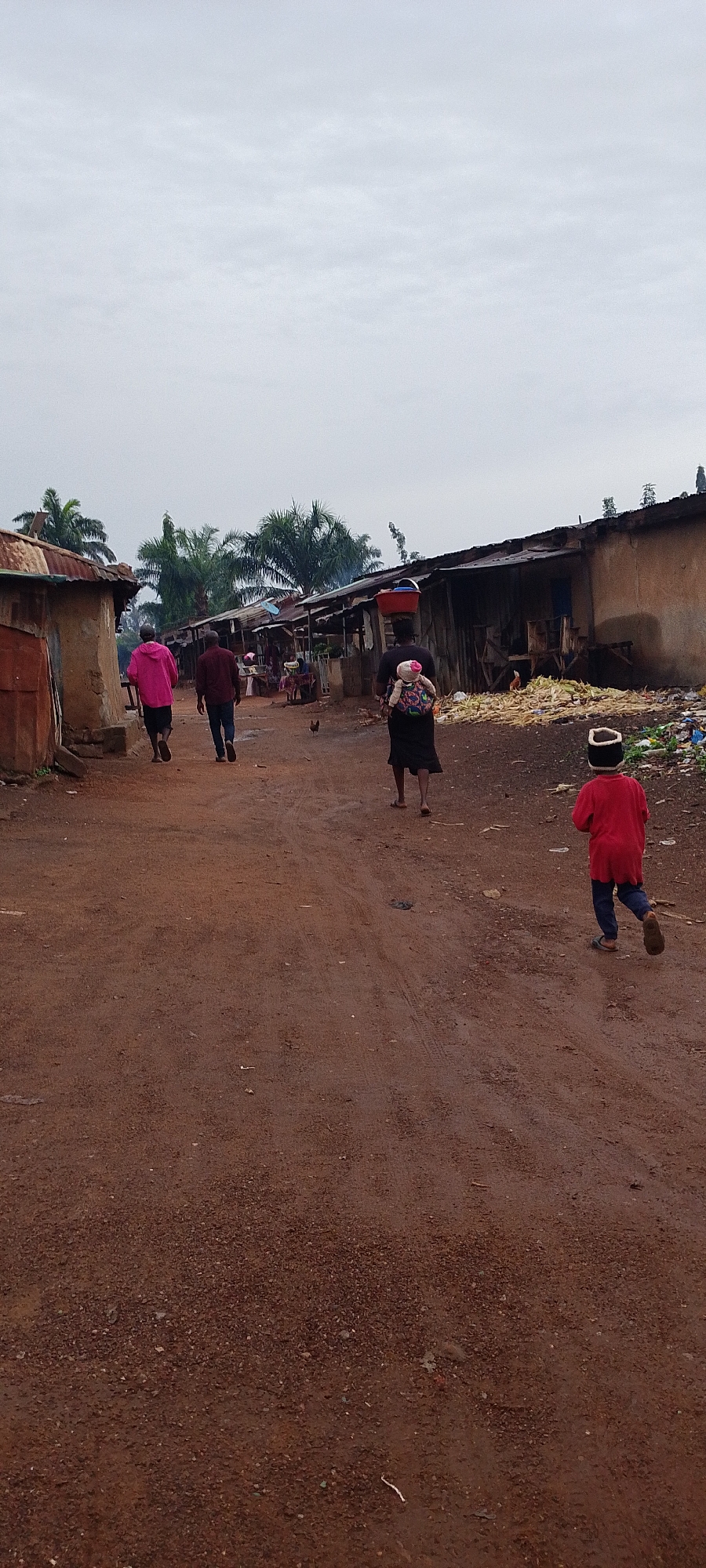 The image depicts a dusty, unpaved street in Gumel, Nigeria, lined with simple, makeshift structures. The sky is overcast. In the foreground, a young child in a red shirt and dark pants runs away from the camera. Further up the street, a person in a black dress with a baby on their back walks away from the camera. To their left, two individuals, one in a pink hooded jacket and the other in a dark shirt and pants, are also walking away. Several chickens are visible on the ground. The structures on either side of the street appear to be made of corrugated metal and mud. Piles of what look like dried corn stalks and refuse are present on the right side of the street. Palm trees are visible in the background.