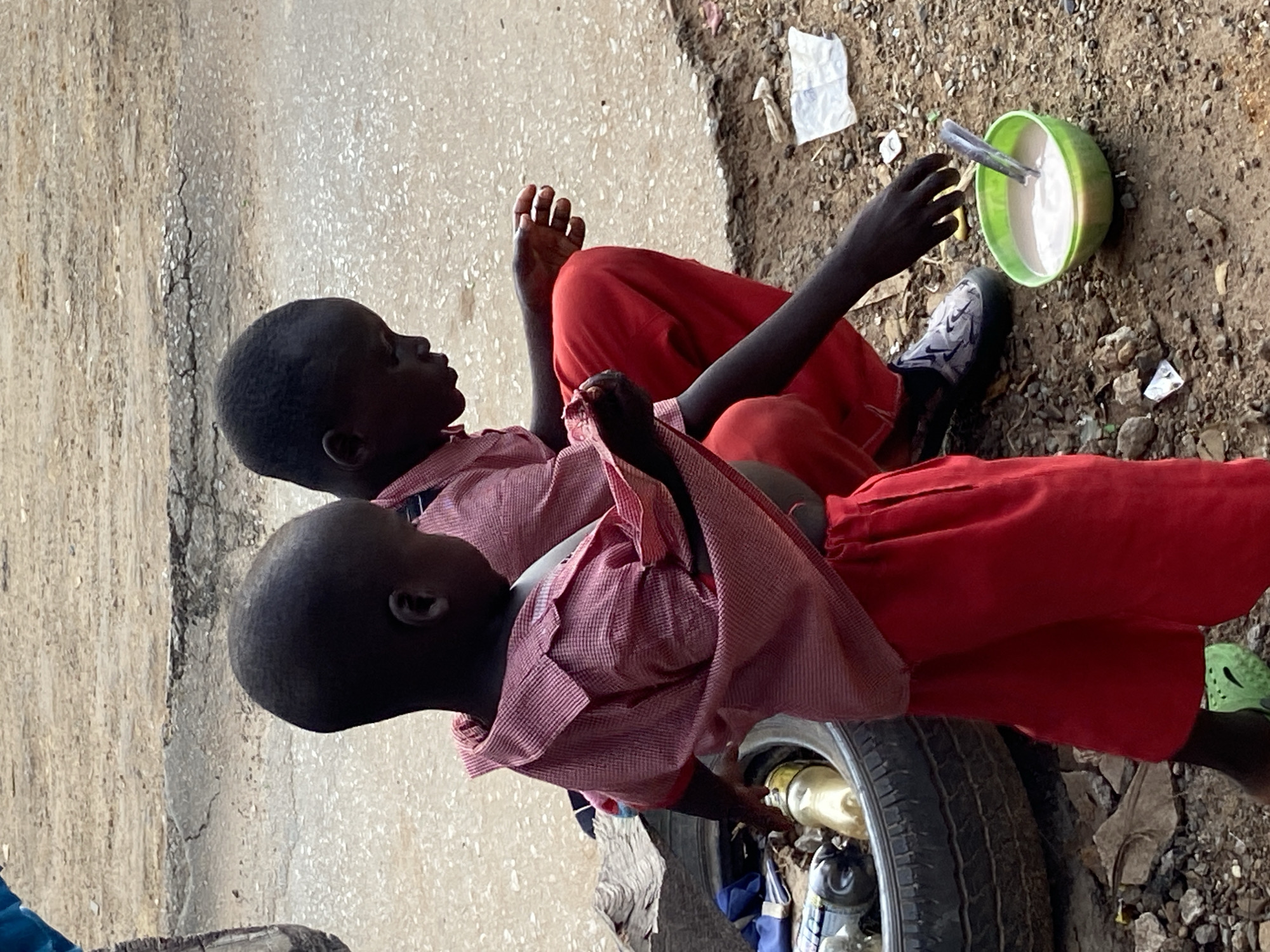 In Mararaba, Nigeria, two young boys in red and white school uniforms are crouched on the dirt ground. The boy on the left, facing away from the camera, is wearing a red top with white checkered patterns and red pants. His left hand is reaching into an old tire that serves as a makeshift container for various items, including bottles and what appears to be trash. The boy on the right, facing towards the left, is wearing a similar uniform and is actively engaged in scooping food from a bright green bowl with a spoon. A second spoon rests in the bowl. Both boys have dark skin and short, dark hair. The environment is a dirt road or pathway with scattered rocks and debris. In the background, a cracked asphalt surface is visible. The overall scene depicts children in a humble setting, highlighting their daily activities and surroundings.