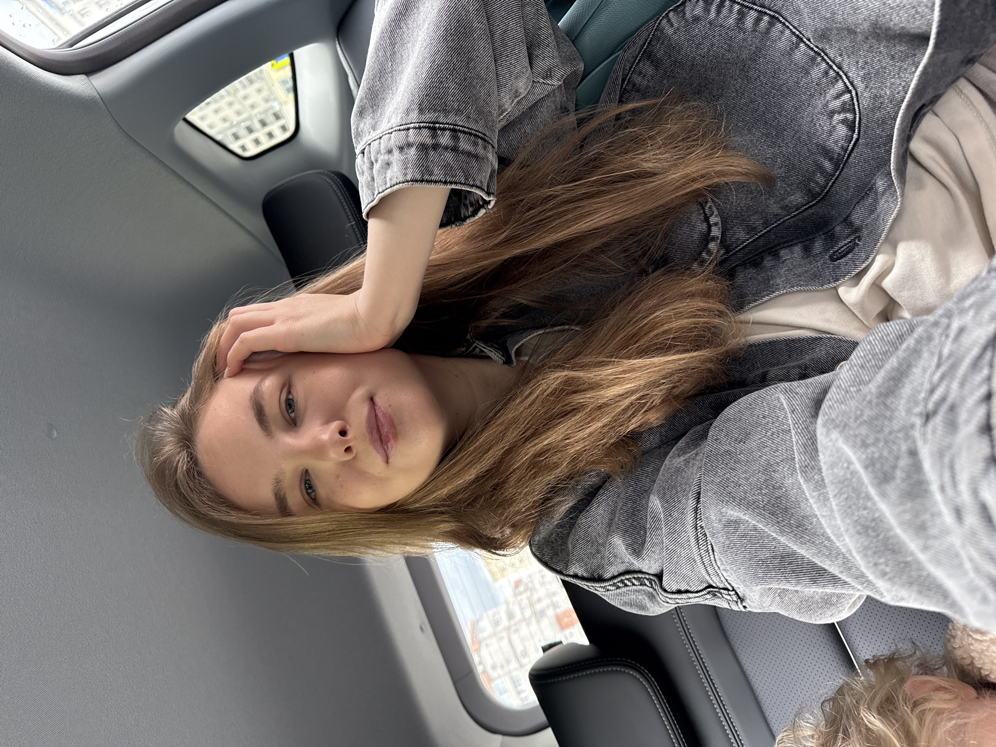 A young woman is captured in a close-up photograph, seemingly a selfie, inside what appears to be a moving vehicle such as a car or train. She has long, light brown hair, fair skin, and blue eyes, looking directly at the camera with a neutral, slightly pensive expression. Her left hand is raised, with fingers resting on her forehead. She is dressed in a dark grey, faded denim jacket, worn over a light-colored (possibly cream or white) top.
The interior of the vehicle is visible, featuring light grey ceiling and pillar coverings, and dark grey or black leather seats with visible stitching. Through the windows, an urban or city landscape can be seen, characterized by multi-story buildings with numerous rectangular windows. The lighting suggests it is daytime, with natural light illuminating the scene. A glimpse of a blue sky is visible through one window, and what may be reflections or raindrops are present on another. In the lower right corner, a portion of another person's head with light, curly hair is visible, indicating the presence of another passenger. The architectural style through the windows is consistent with European or post-Soviet cities, aligning with the provided context of Vladimir, Russia. There is no visible text in the image.