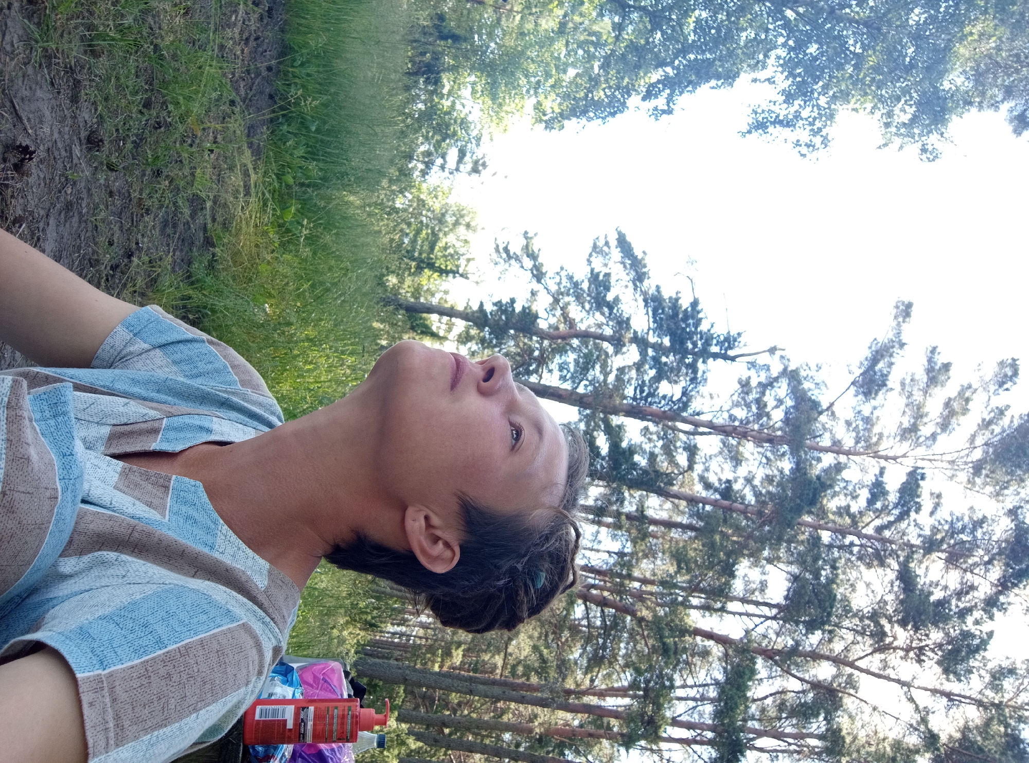 The image shows a person with short, dark hair styled upwards, looking up and to the left. They are wearing a striped shirt with blue and tan horizontal bands. The background is a forest with tall trees and green grass. The lighting suggests it is daytime. The location is identified as Podborov'e, Russia, indicating an outdoor, likely rural or park-like setting. There are no other people visible, and the person appears to be alone. In the background, on a surface that could be a table, are two bottles, one red and one blue. The overall scene depicts a person enjoying nature.